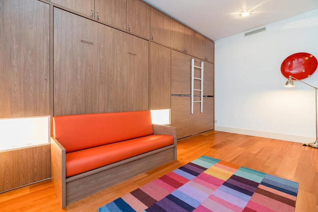 Una colorida sala de estar tiene almacenamiento de madera, asientos naranjas y una alfombra estampada.