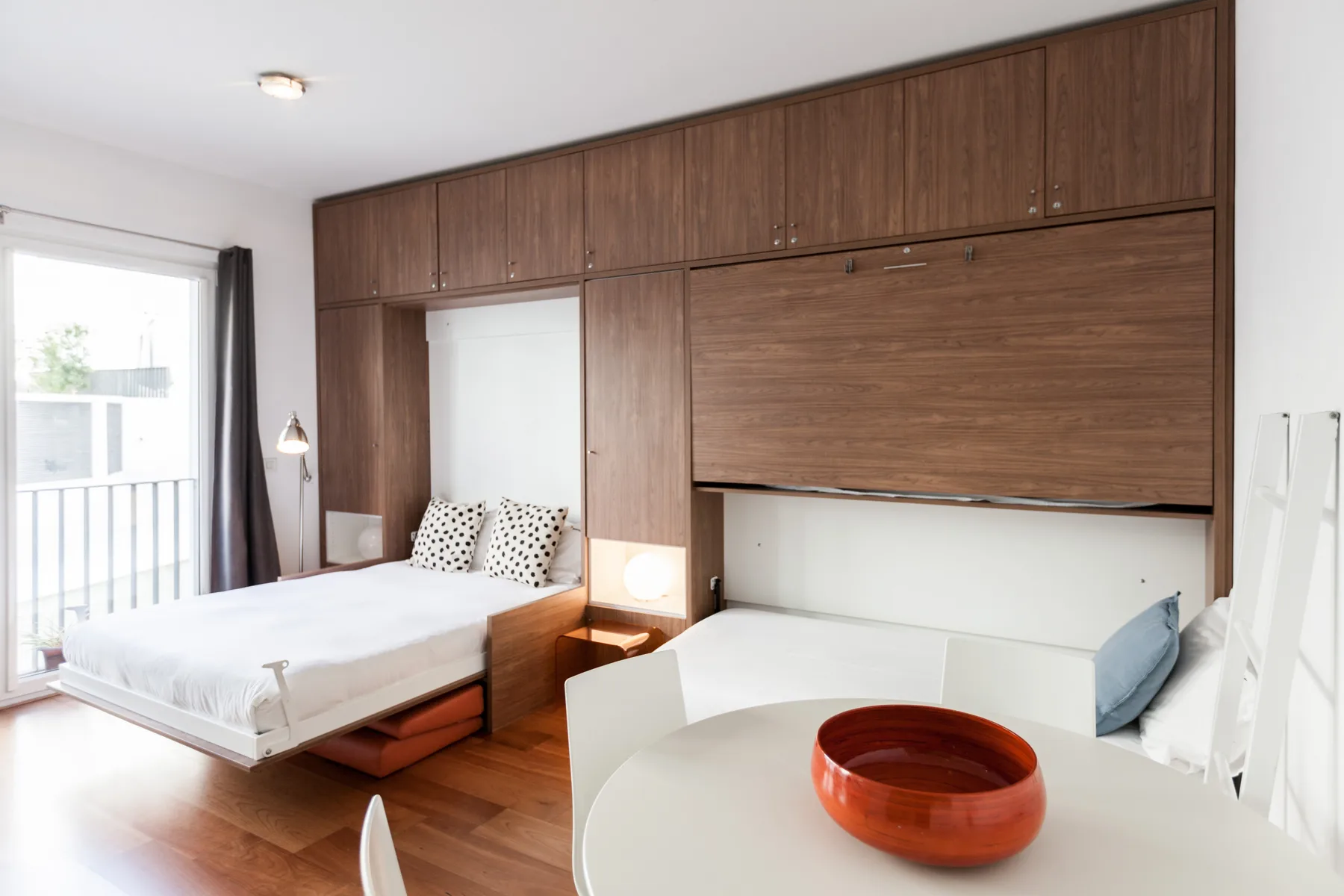 Un dormitorio luminoso con zona de comedor, cama, mesa y armarios de madera.