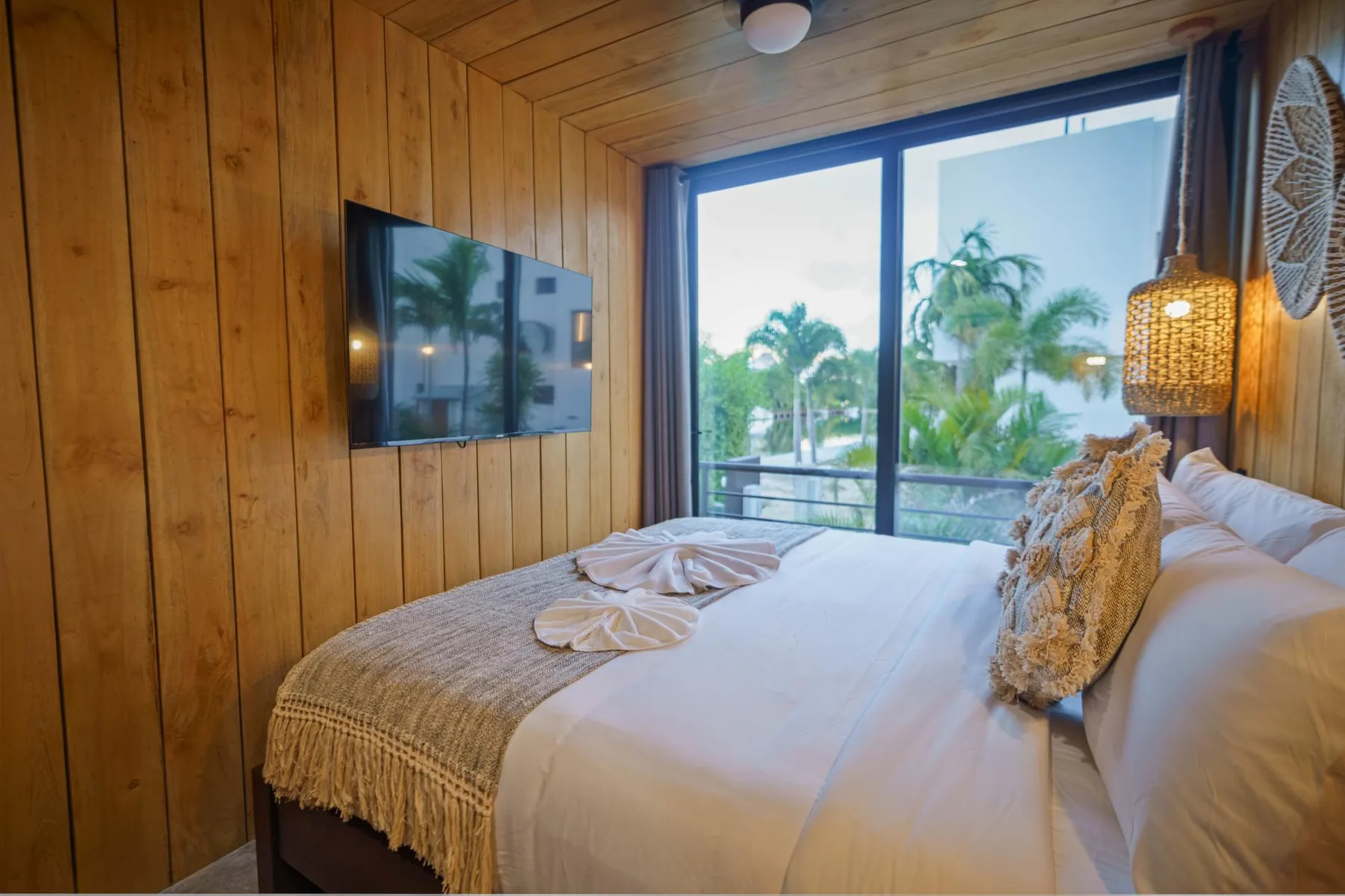 A bedroom with a bed beside a large window overlooking palm trees.