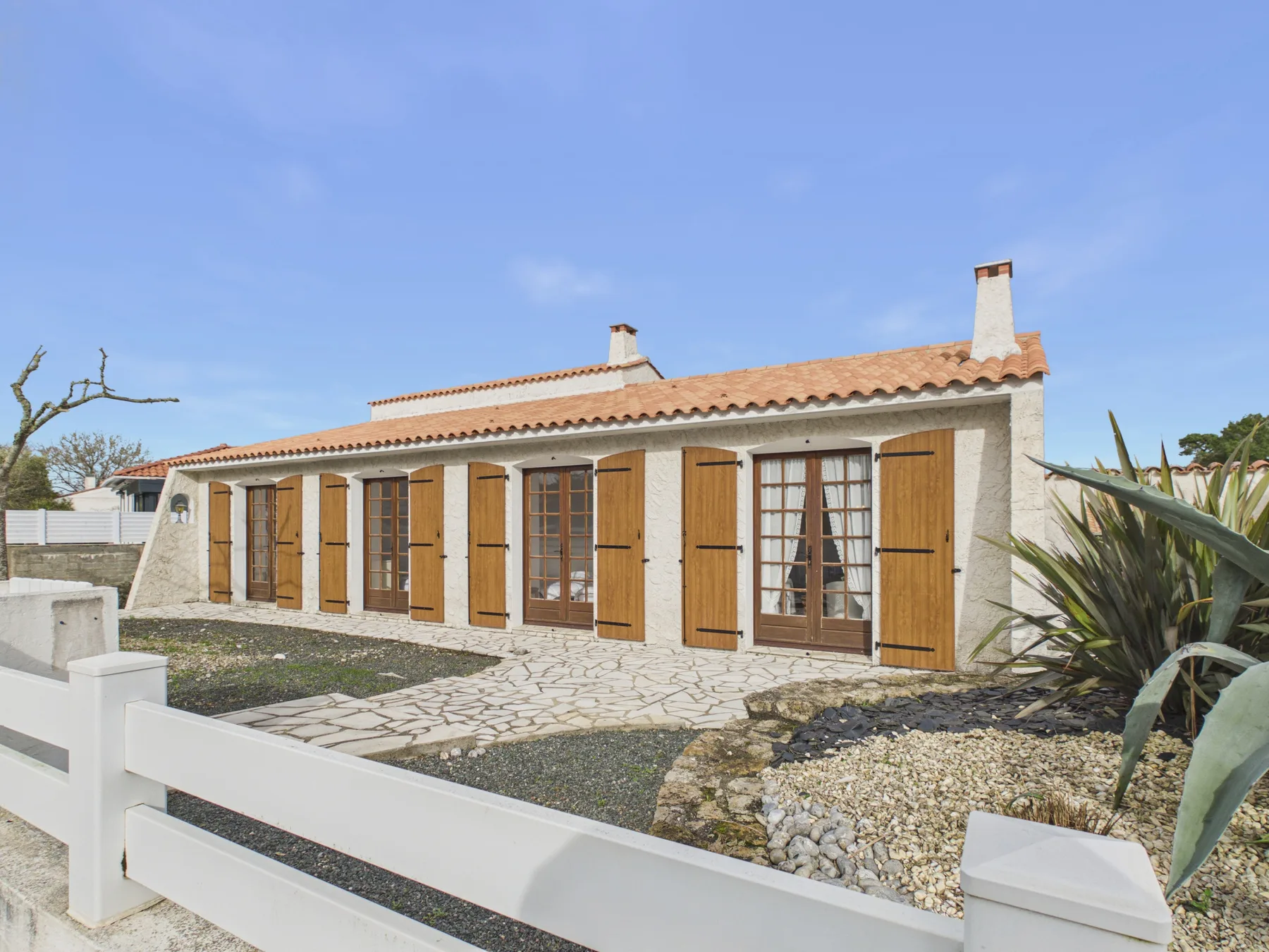 A single-story house with wooden shutters sits behind a white fence.