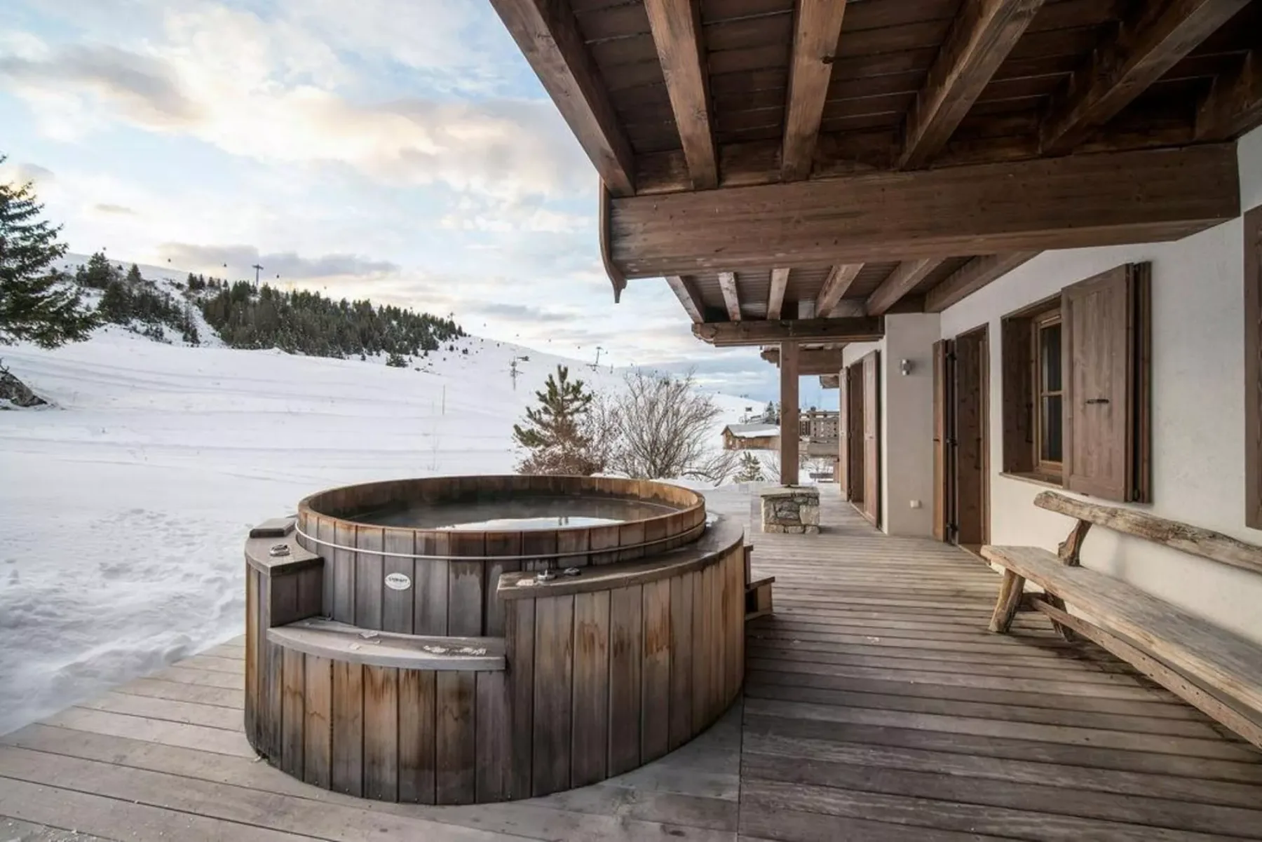 Una bañera de hidromasaje de madera está en una terraza junto a un edificio, con una ladera nevada al fondo.