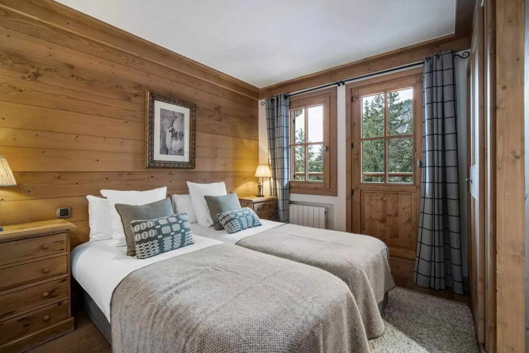 A bedroom with two beds, wood-paneled walls, and windows overlooking trees.