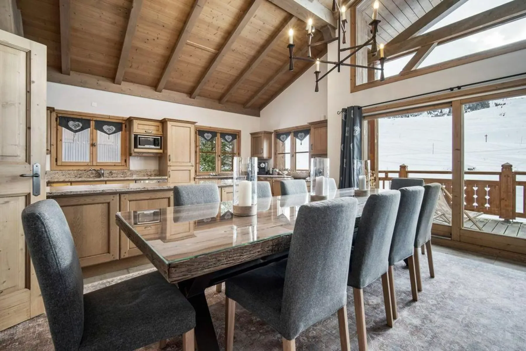 A dining area with a long table and kitchen cabinets beneath a wood-beamed ceiling.