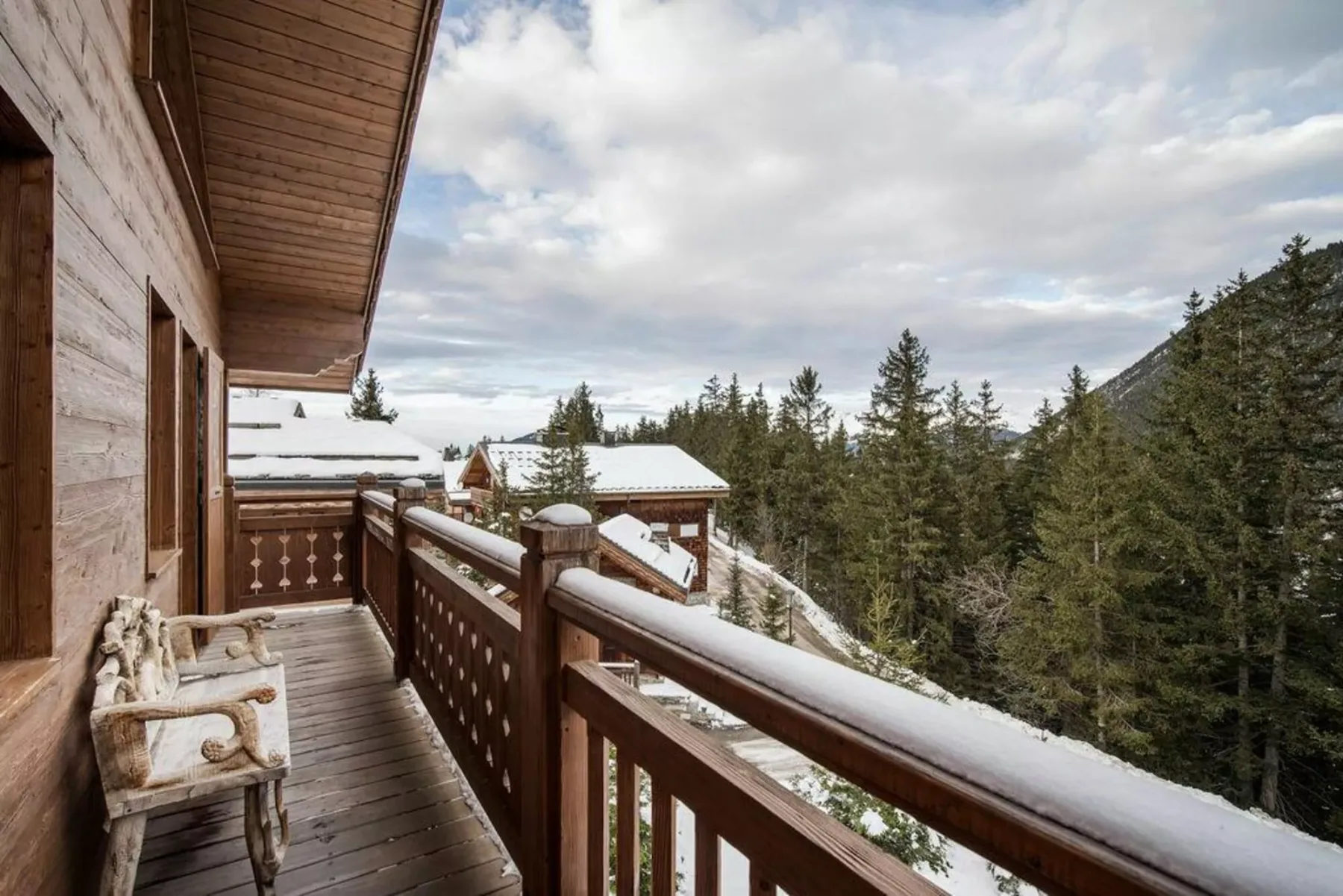 A wooden balcony overlooks snow-covered rooftops and a forested hillside.