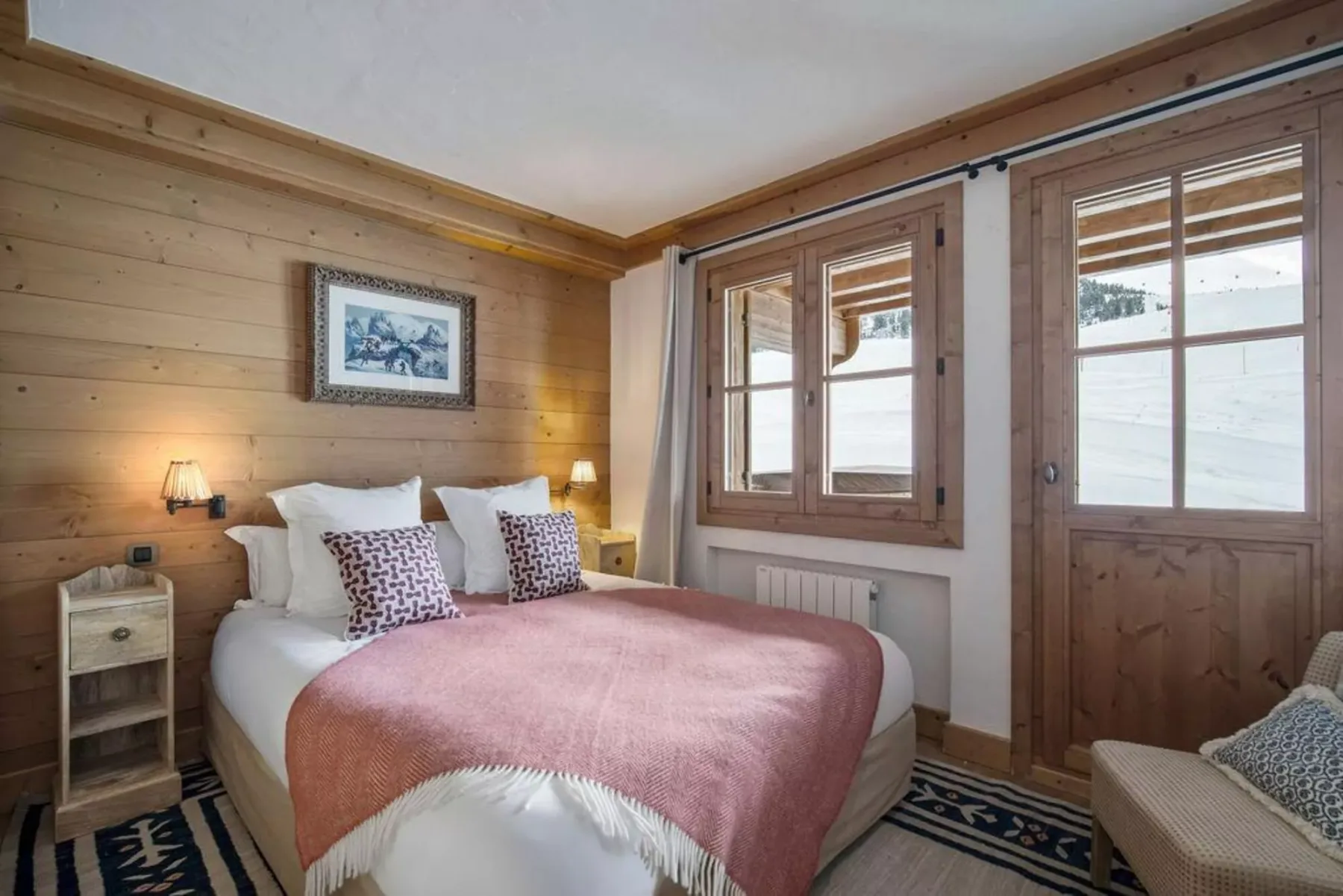 A bedroom with a bed covered by a rose-colored blanket and wooden wall paneling.