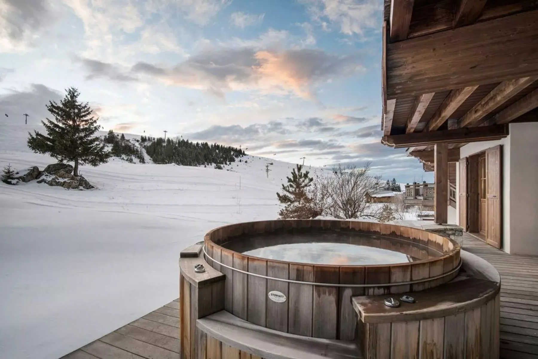 A wooden outdoor hot tub sits beside a building overlooking a snowy landscape.