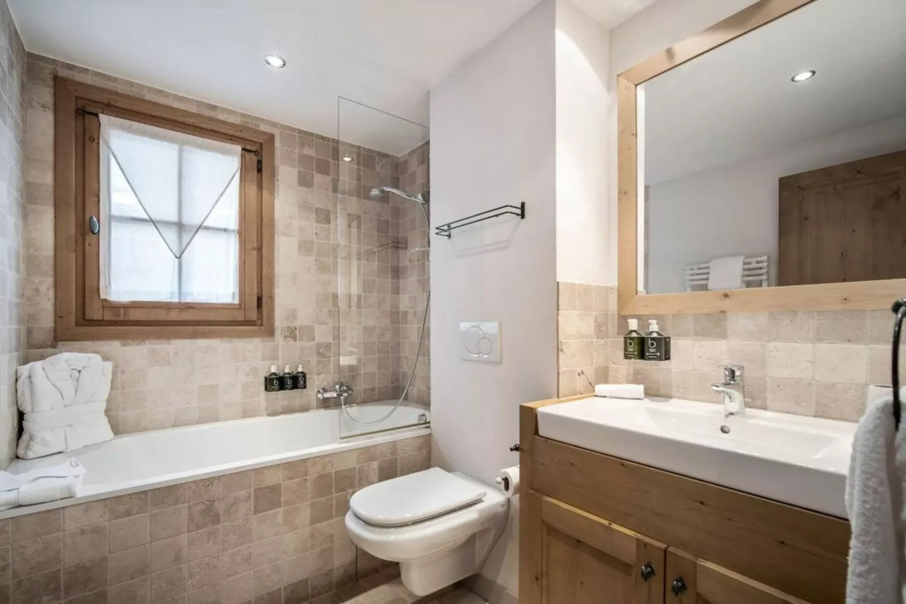 A tiled bathroom with a bathtub, wall-mounted toilet, and wooden vanity.