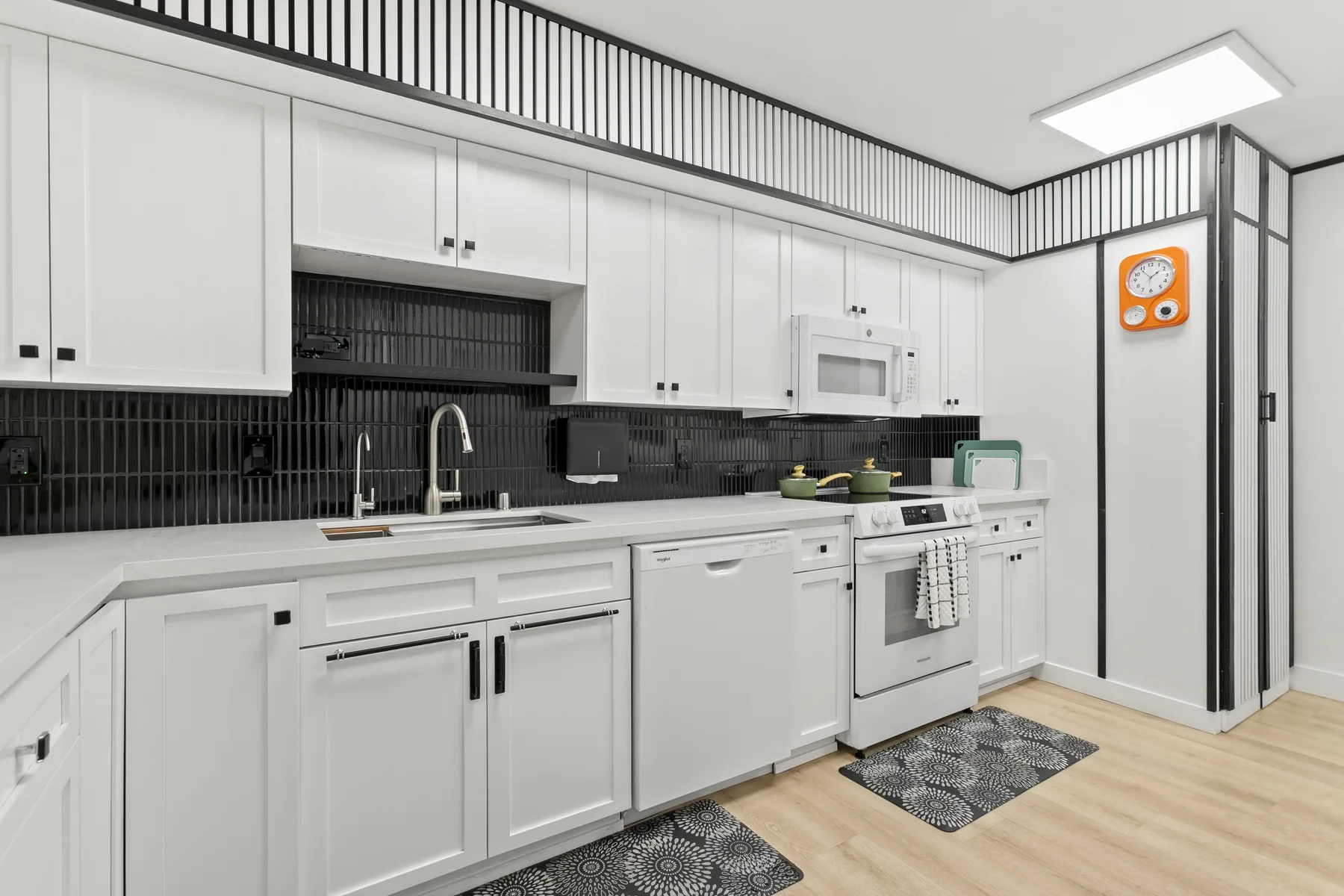 Una cocina moderna y luminosa con gabinetes blancos, salpicadero de azulejos oscuros y suelo de madera clara.