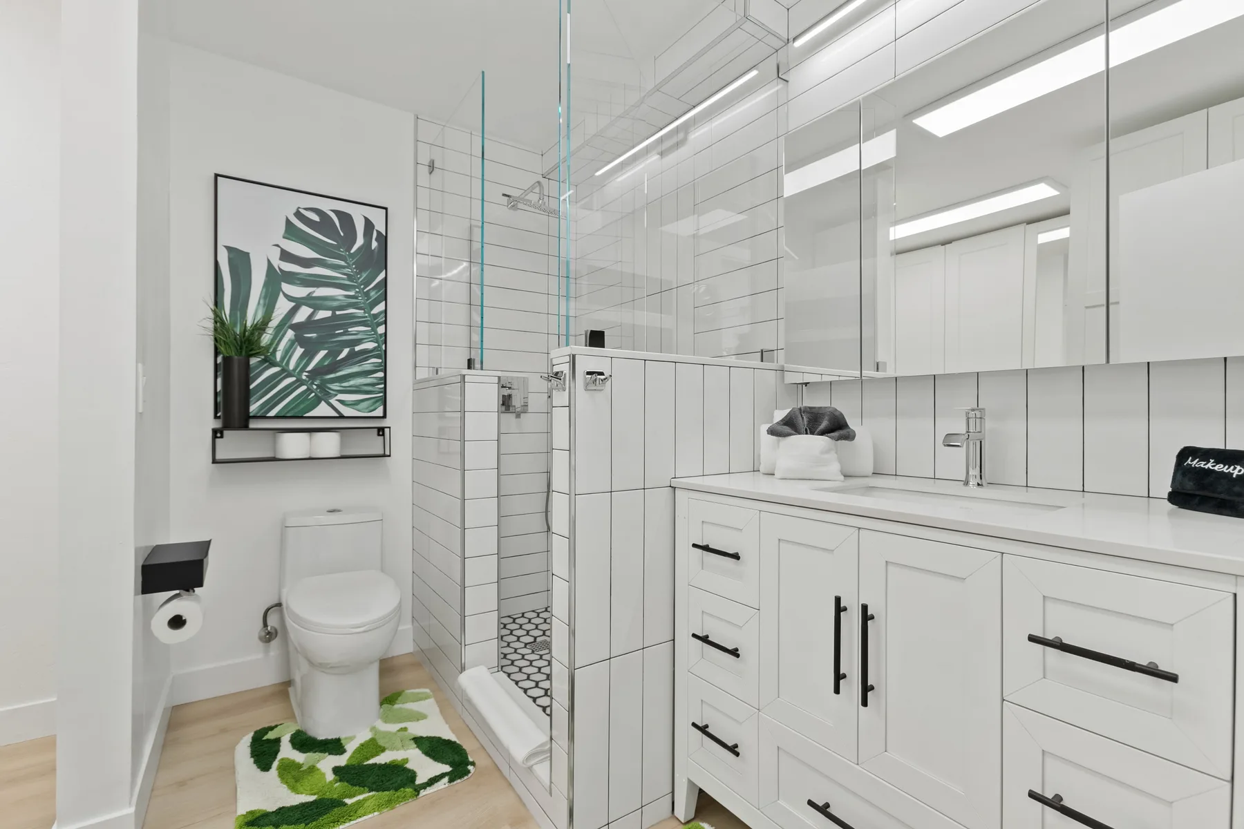 Un baño luminoso con ducha de vidrio, tocador blanco y arte vegetal en la pared.