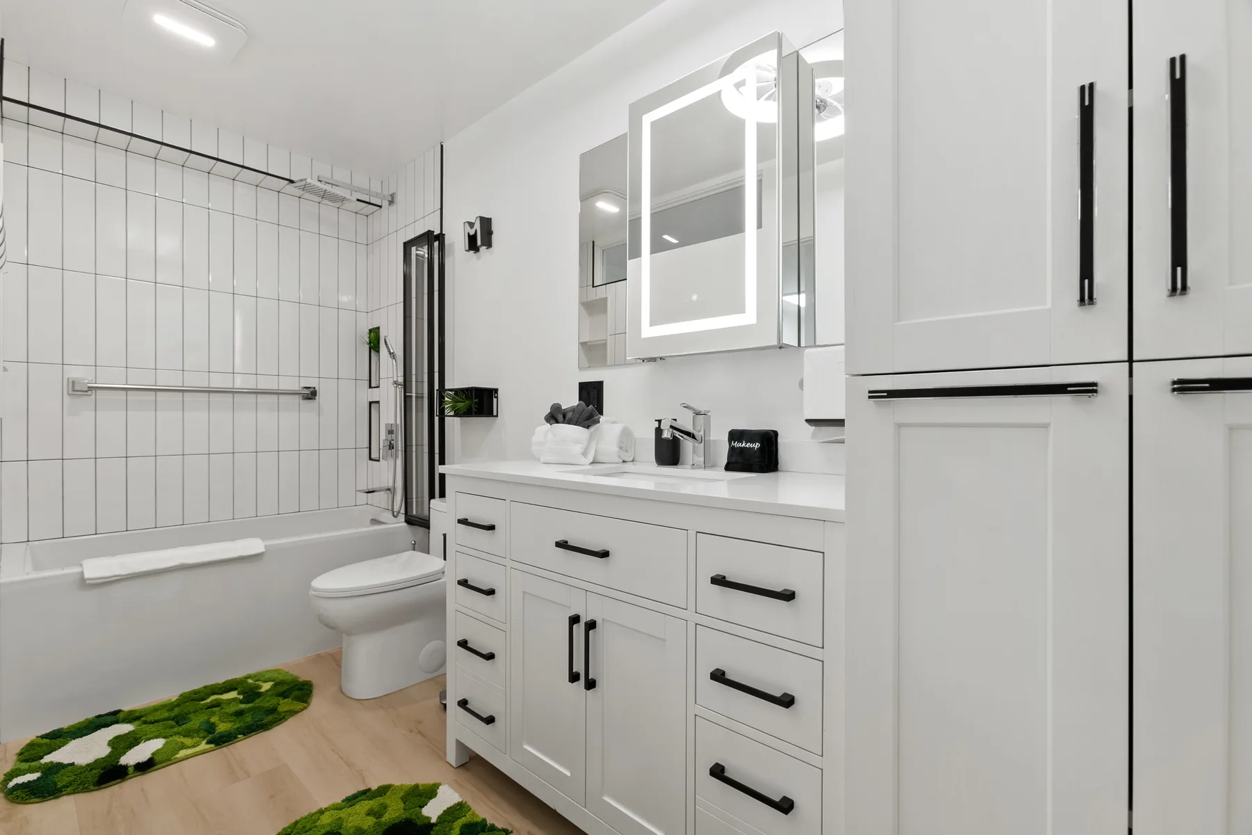 Un baño luminoso con ducha de azulejos, lavabo y gabinetes blancos.
