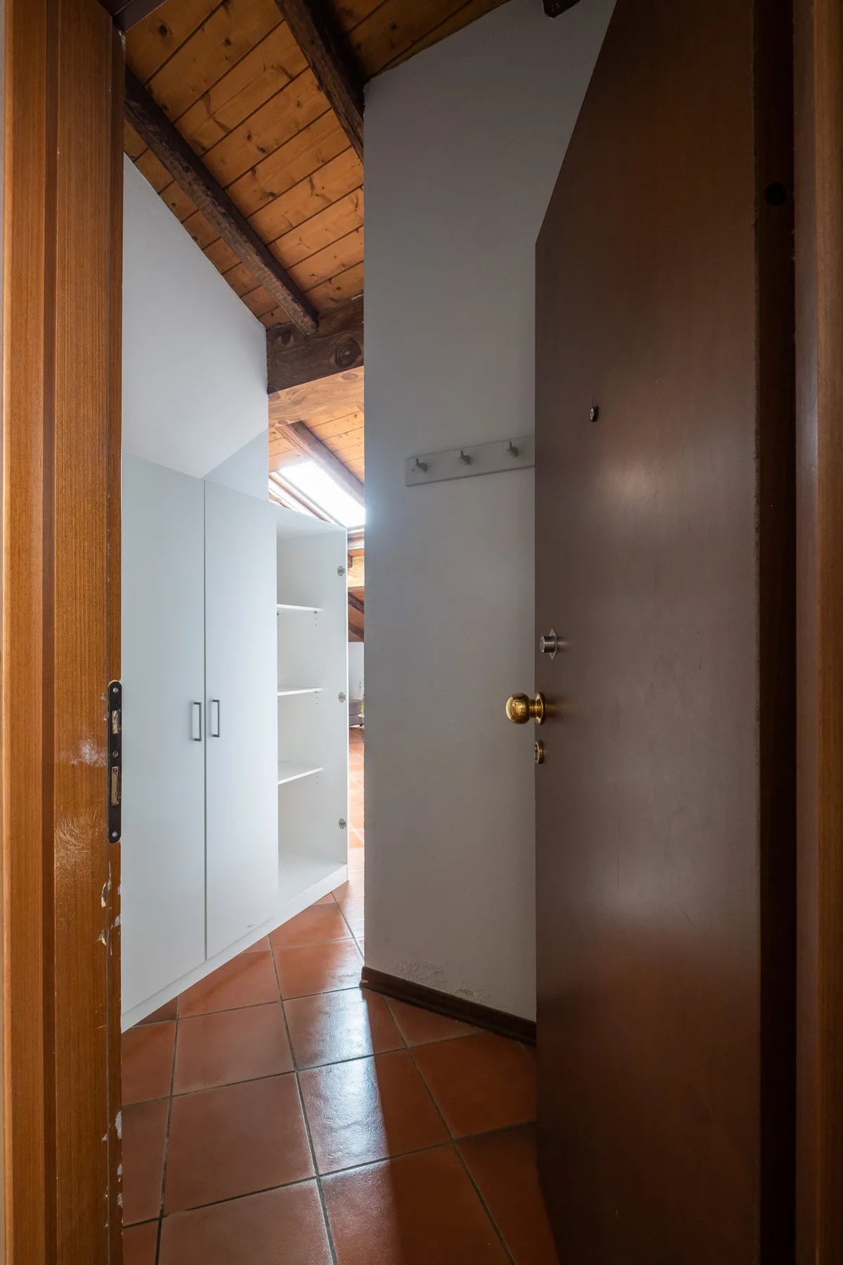 Un pasillo con suelo de baldosas pasa junto a armarios blancos bajo un techo inclinado de madera.