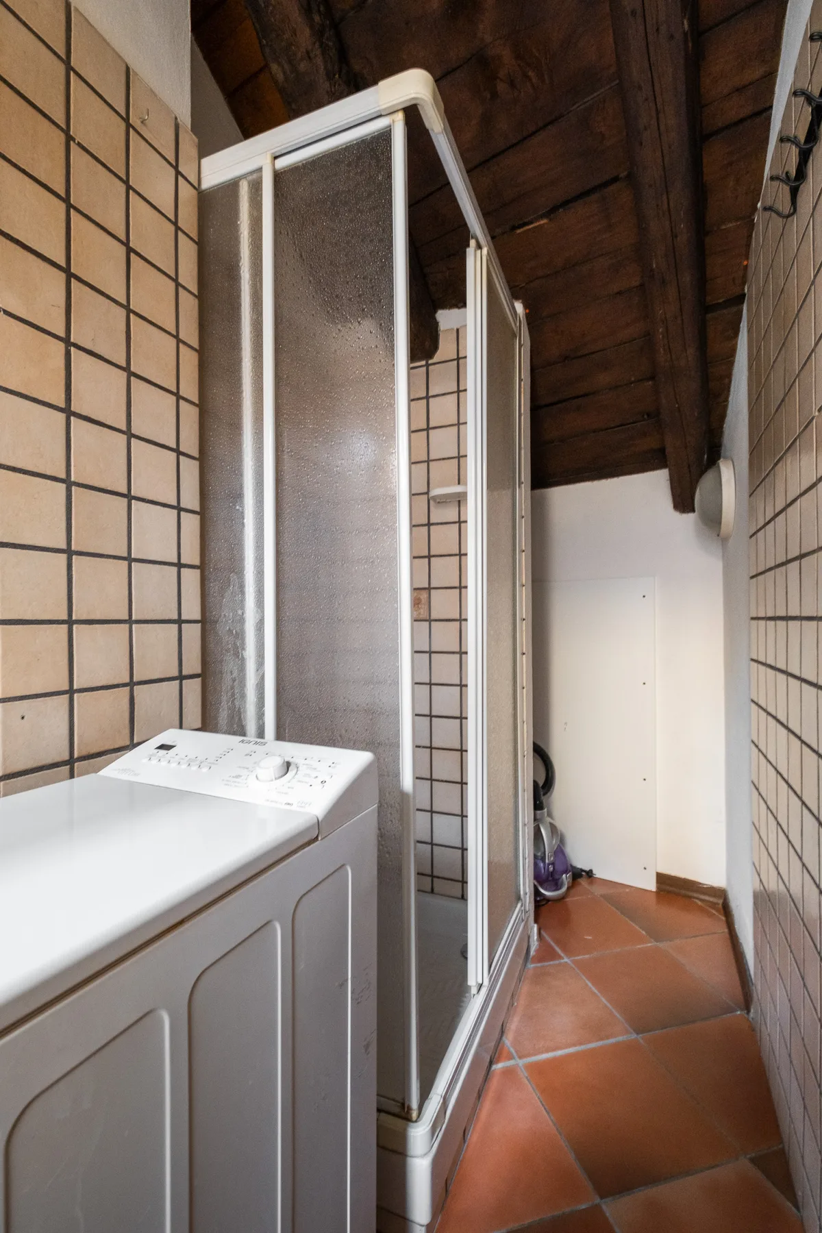 Un baño estrecho con ducha, lavadora y techo de madera inclinado.