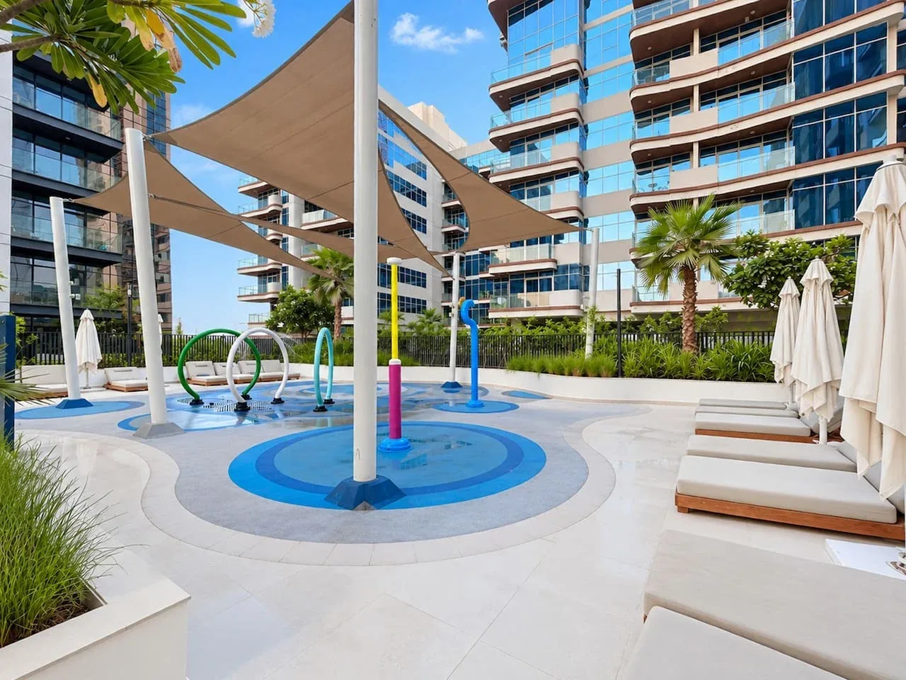 A sunny outdoor pool area with blue circular water features and shade sails.