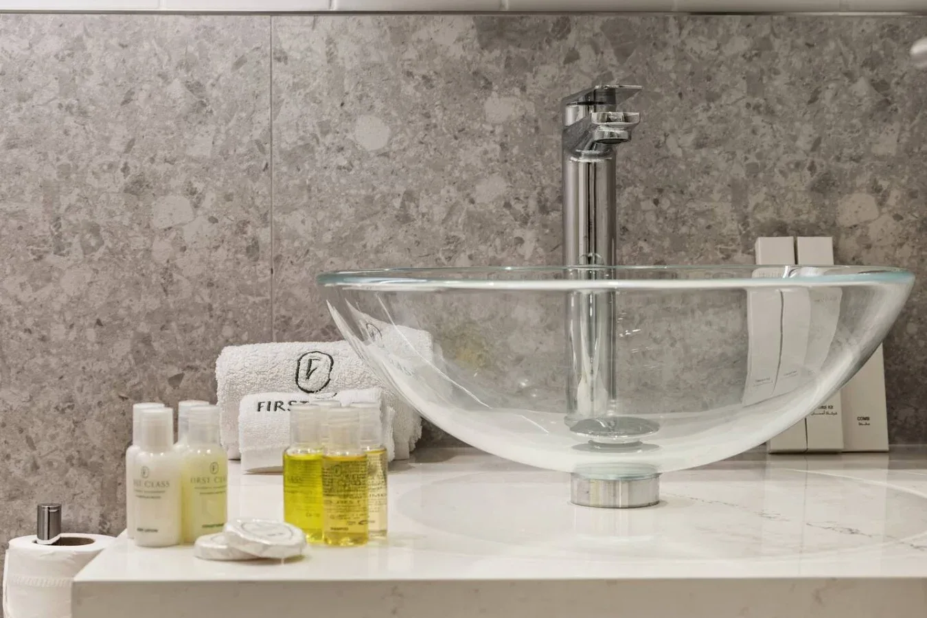 A bathroom vanity features a clear vessel sink, chrome faucet, and toiletries.
