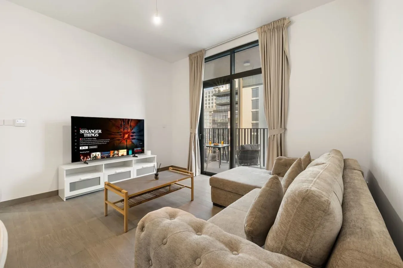 A living room with a television, seating, and tall curtained windows opening onto a balcony.
