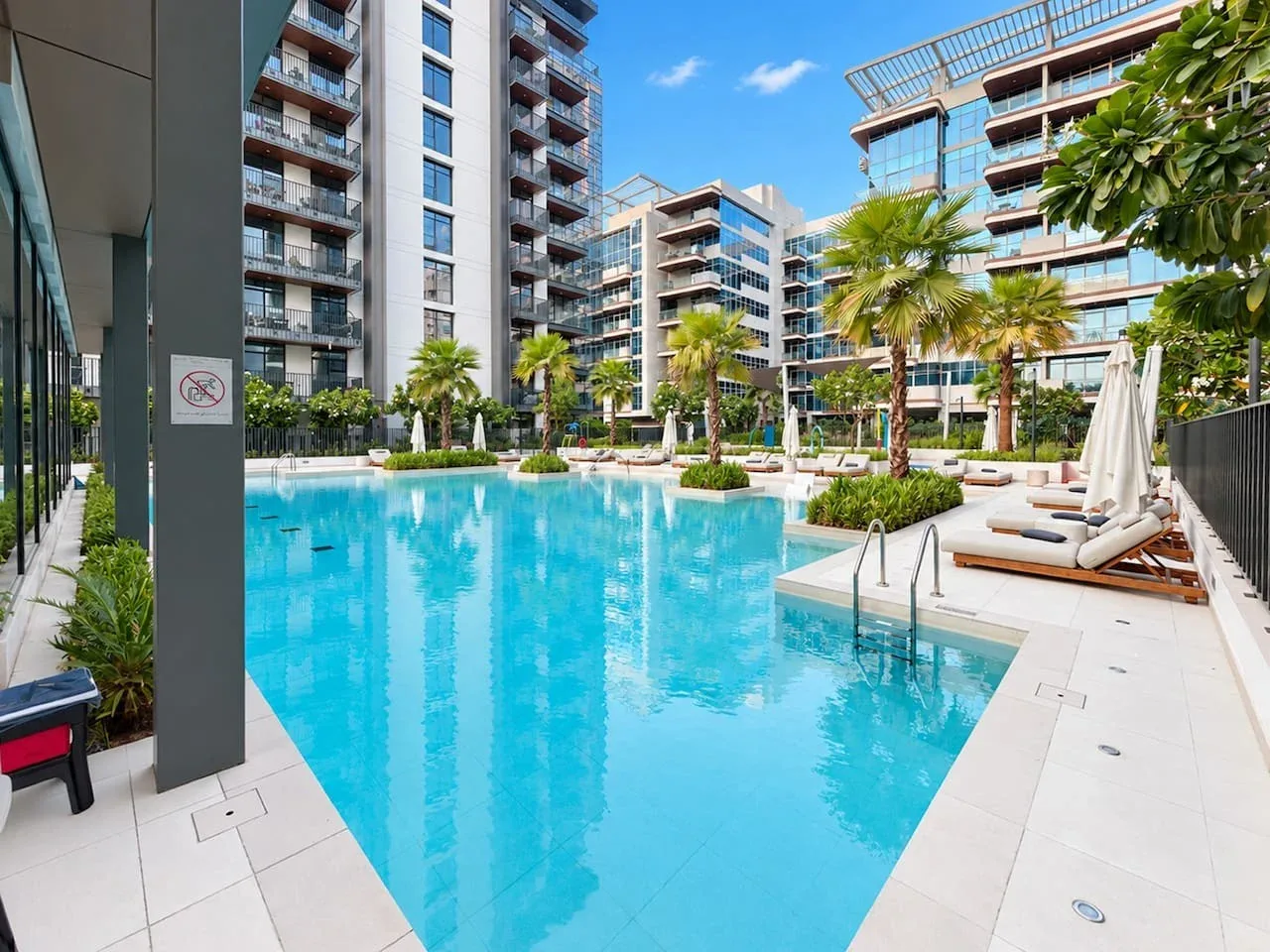 A swimming pool surrounded by apartment buildings, palm trees, and lounge chairs.