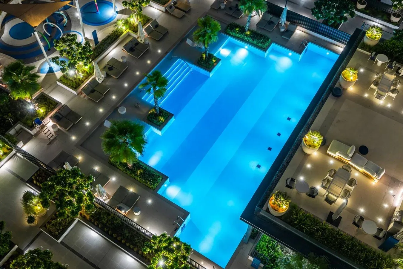An illuminated swimming pool is surrounded by lounge seating and landscaped paths.