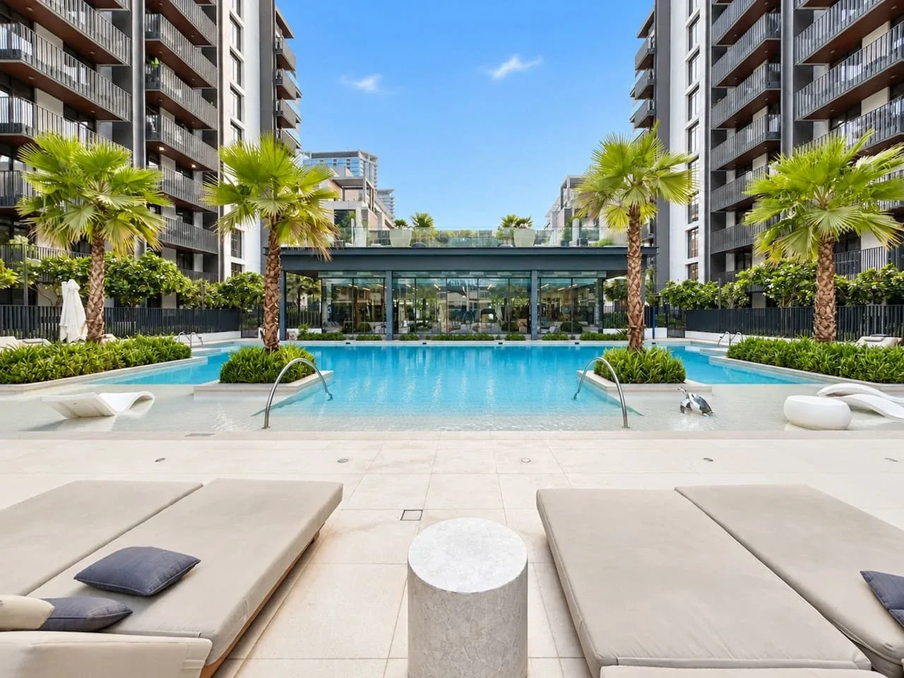 An outdoor swimming pool sits between modern apartment buildings, with loungers in the foreground.