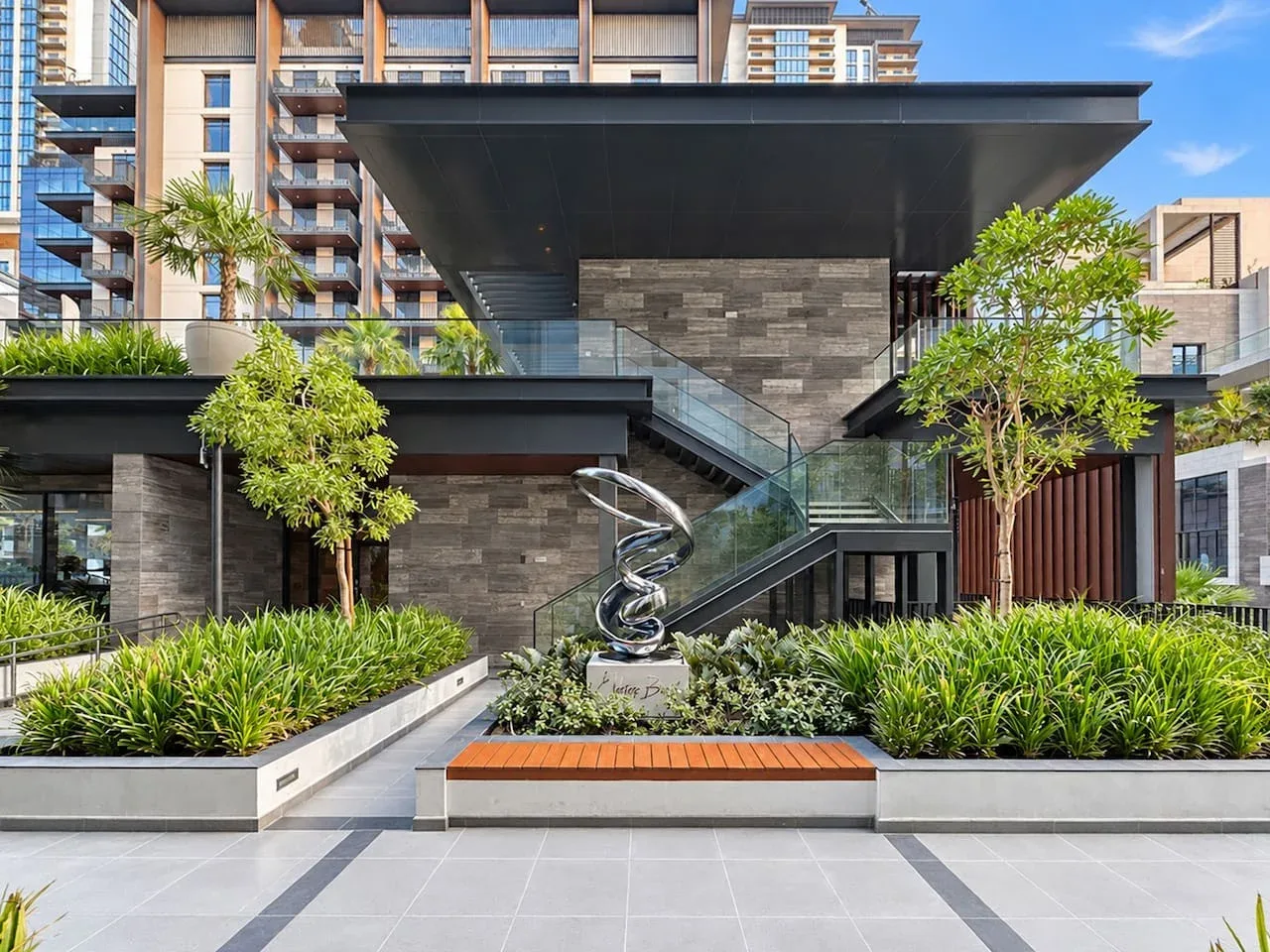 A modern building entrance opens onto a landscaped courtyard with a sculpture.