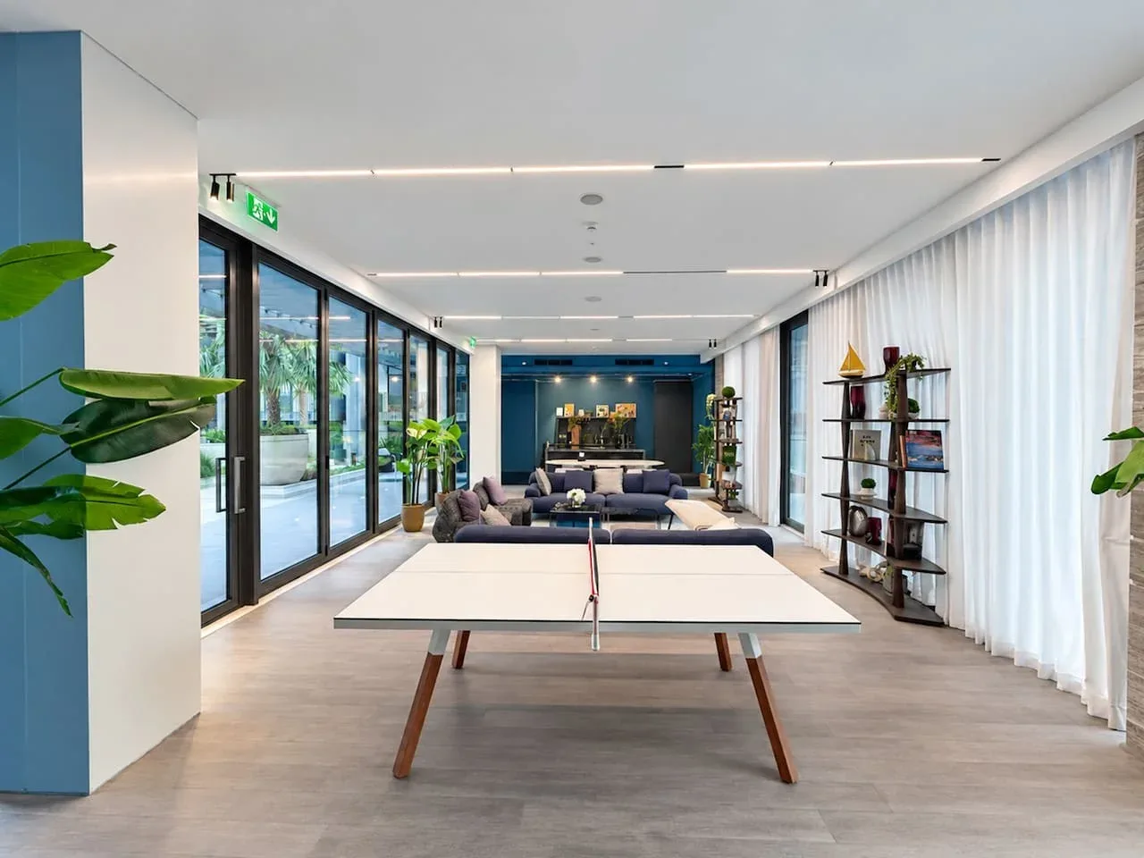 A bright lounge with a long table in front of a seating area and large windows.