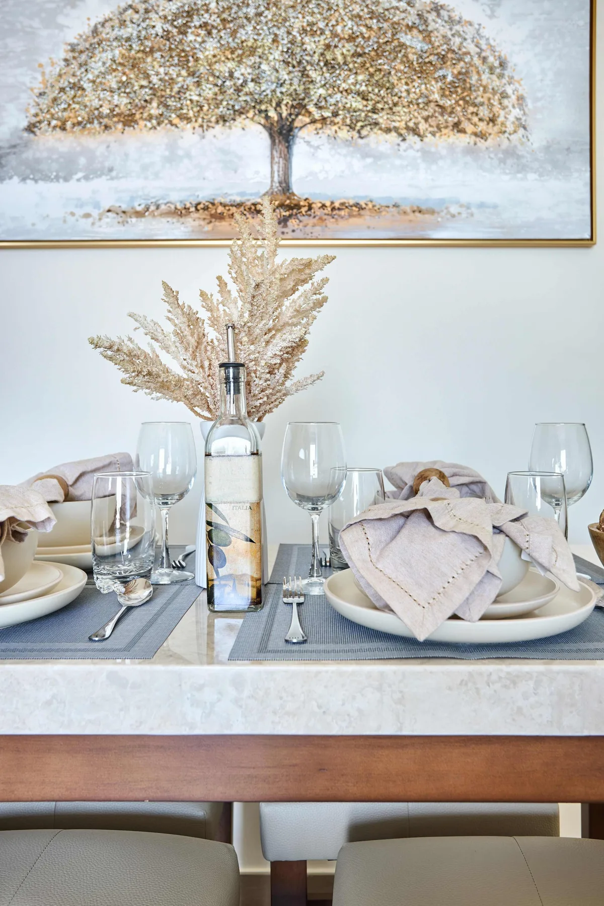 Una mesa de comedor está puesta con platos, cristalería y una botella bajo un cuadro de un árbol.