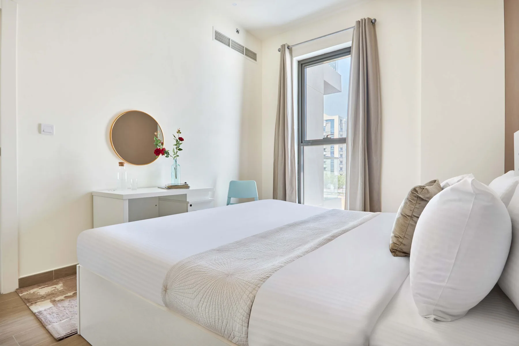 Un dormitorio luminoso con una cama tendida, un tocador y una ventana alta.