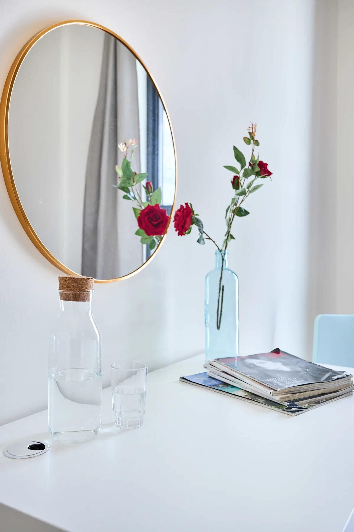 Una mesa blanca con un espejo, flores en jarrones de vidrio y revistas apiladas.