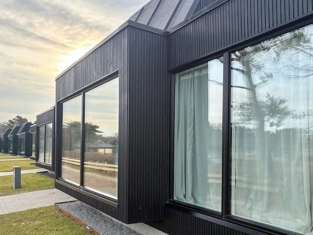 Una casa moderna de revestimiento oscuro y grandes ventanas junto a un camino pavimentado.
