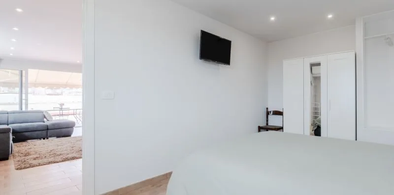 A bright bedroom with a wall-mounted television and an open view into the living area.