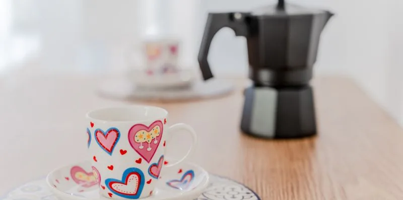 A coffee maker and decorated cup sit on a wooden table.