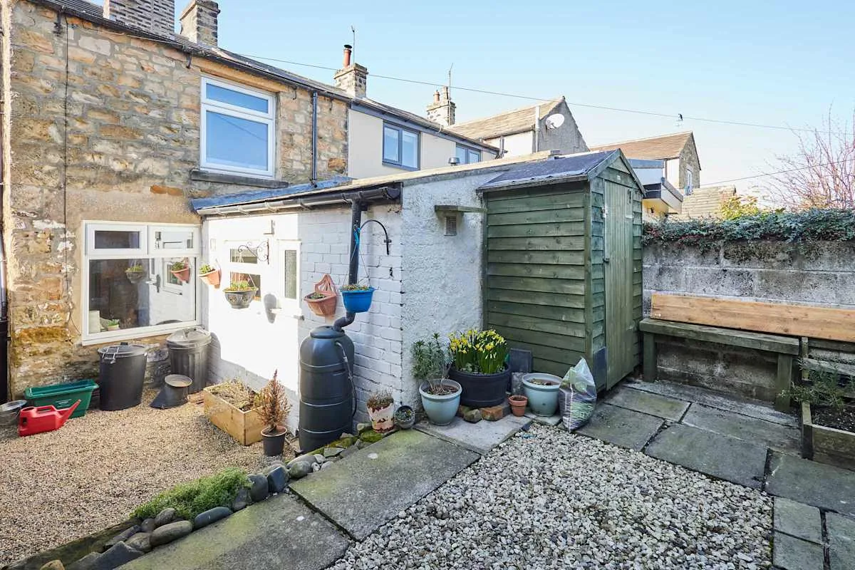A gravel courtyard with a green shed, potted plants, and a wooden bench.
