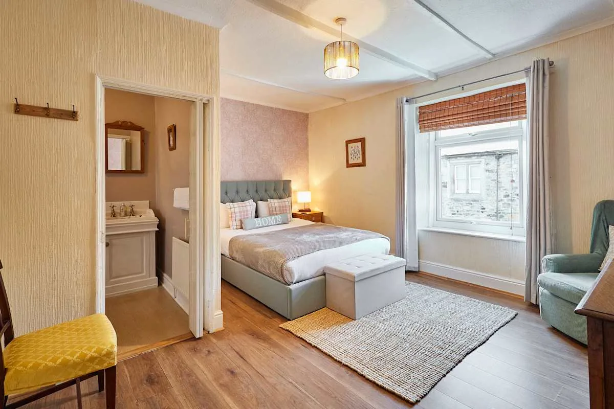 A bedroom with a neatly made bed, an adjoining vanity area, and a large window.