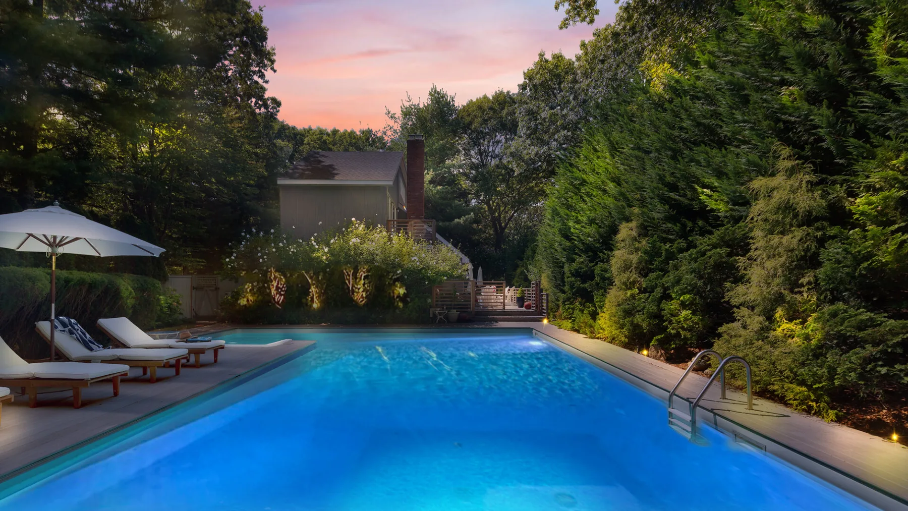 An outdoor swimming pool sits among leafy trees beneath a pink evening sky.
