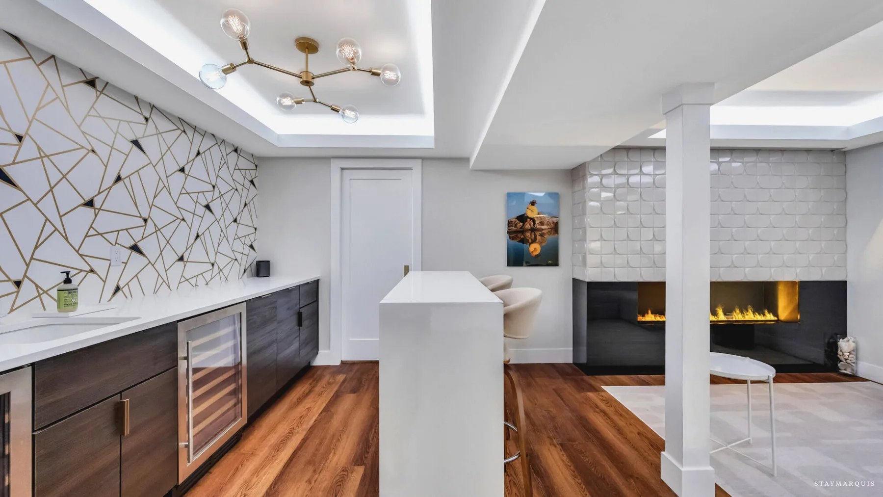 A modern kitchen opens onto a living area with a fireplace and wood flooring.