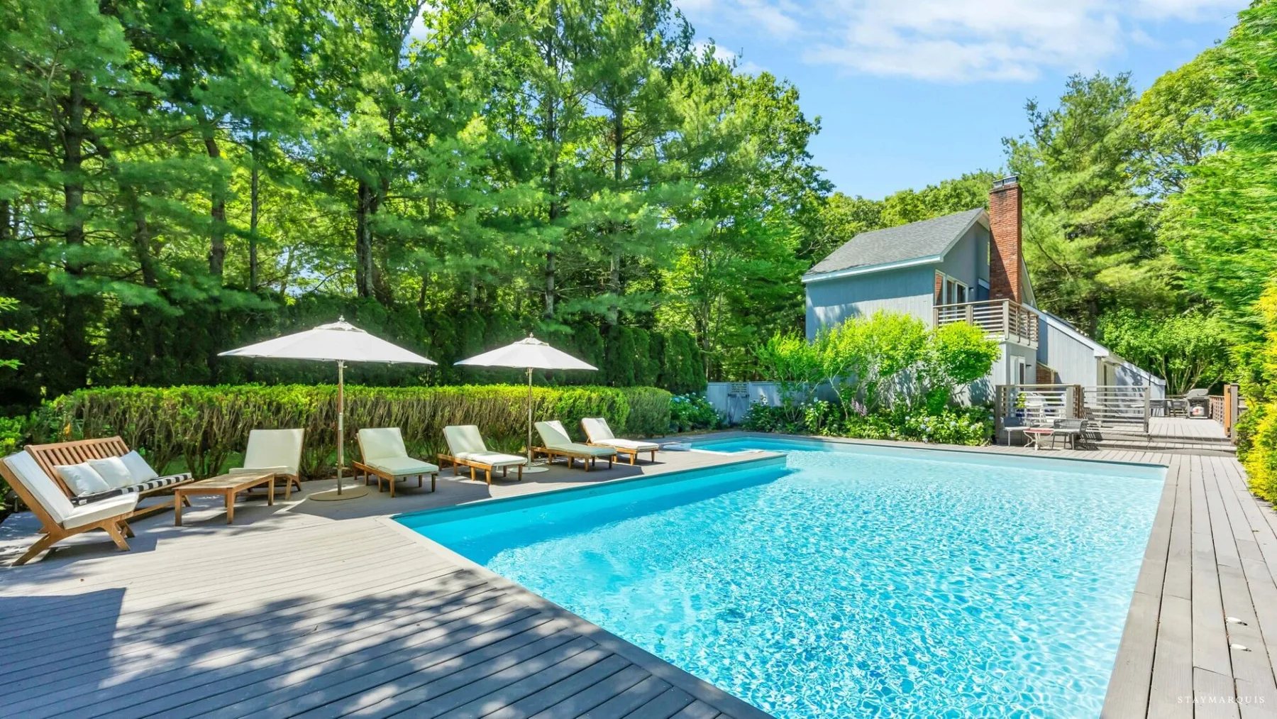 An outdoor swimming pool is surrounded by a wooden deck, lounge seating, and trees.