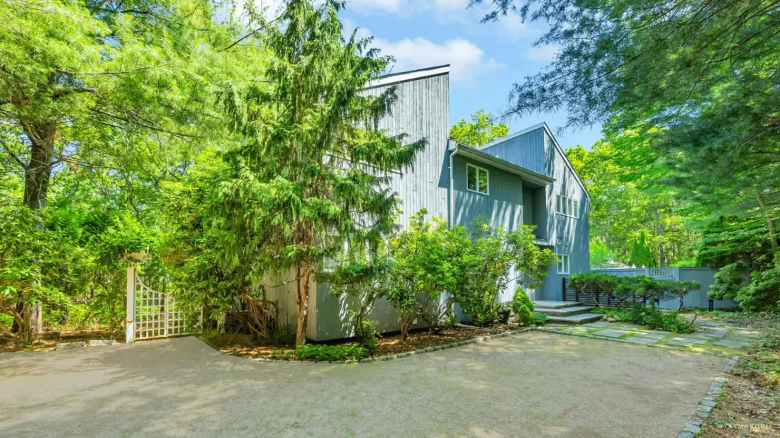 A modern house stands among leafy trees beside a paved driveway.