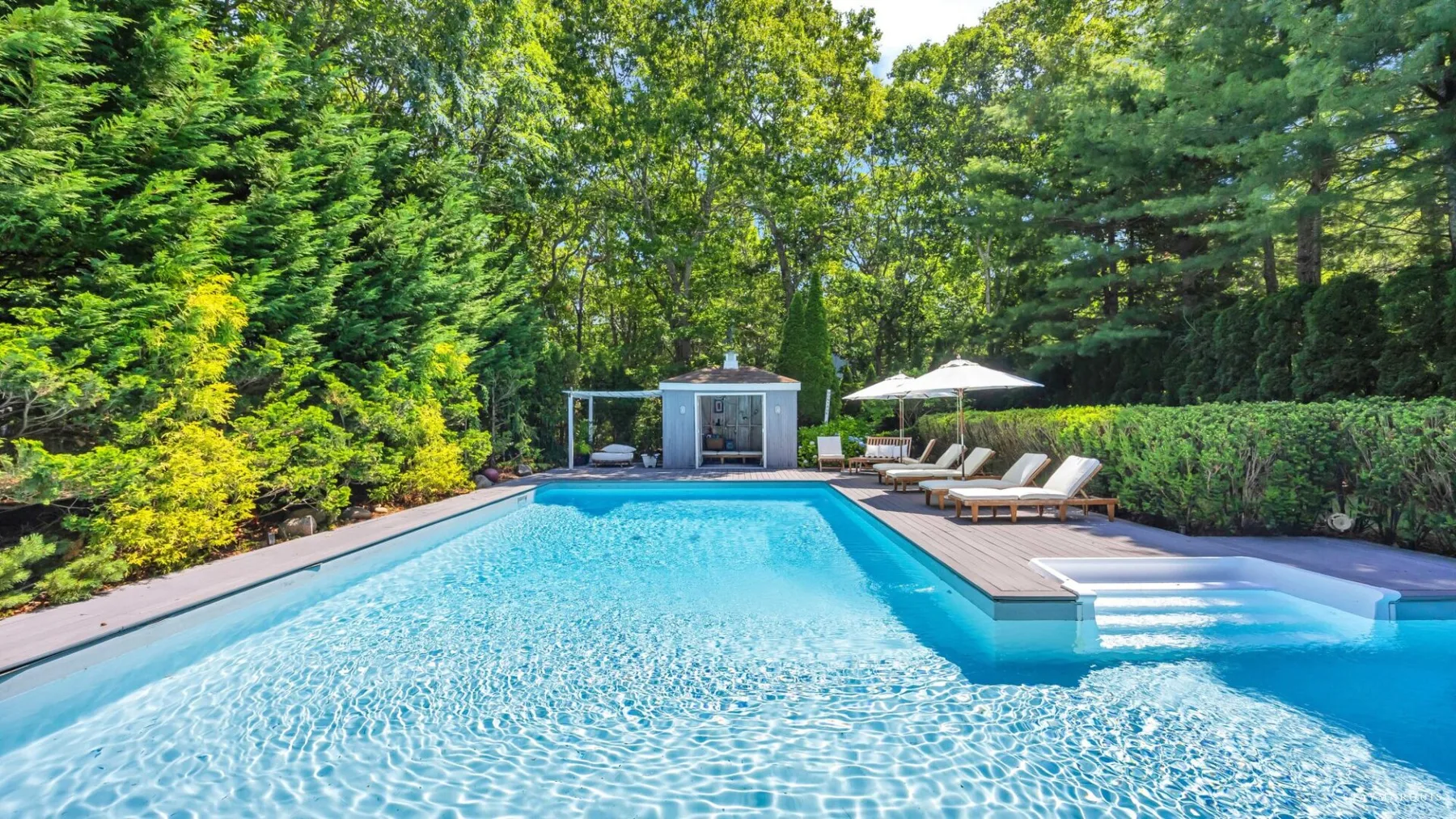 A swimming pool with a wooden deck and lounge seating sits among green trees.