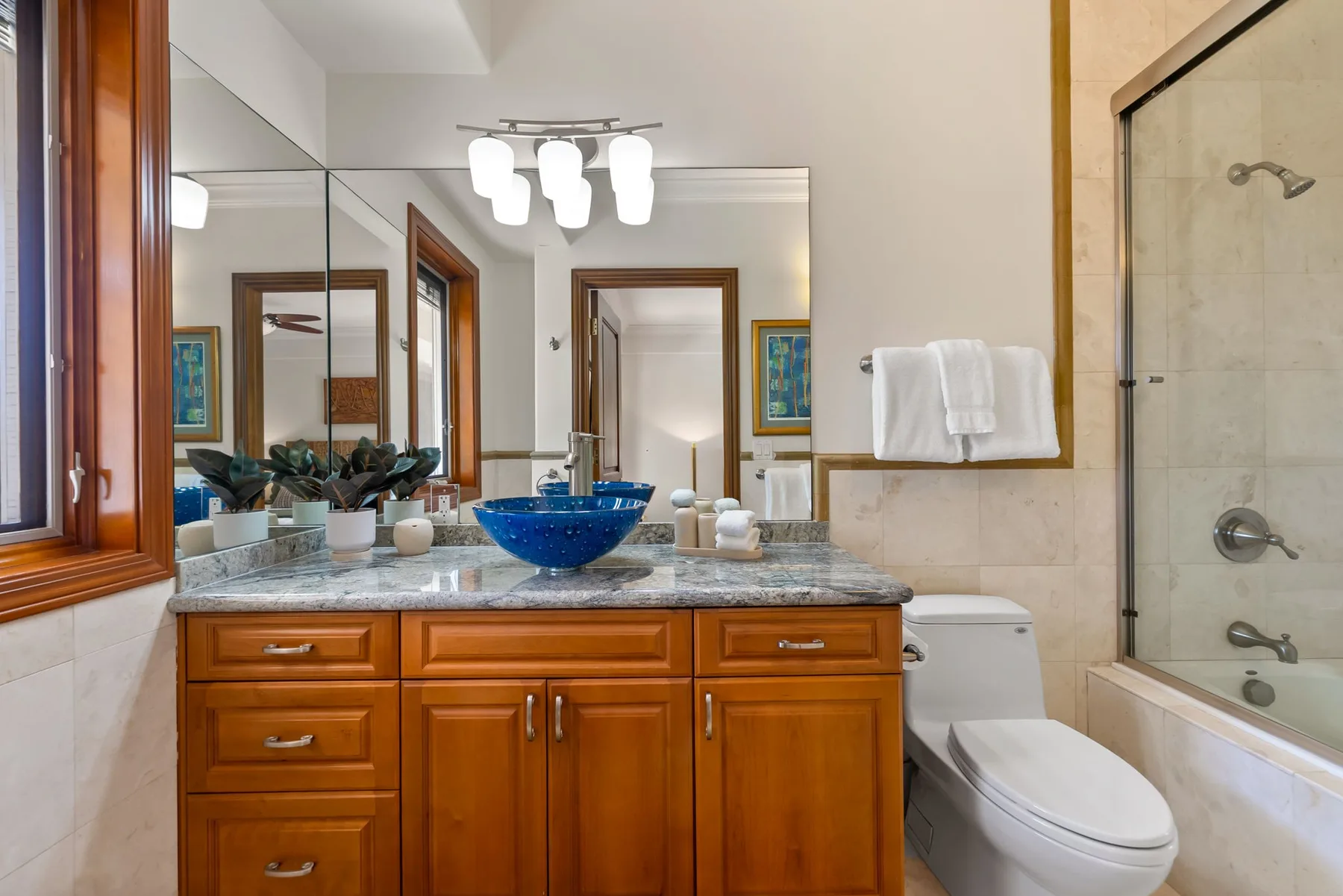 Un baño con mueble de madera, lavabo azul sobre encimera, inodoro y bañera con mampara de vidrio.