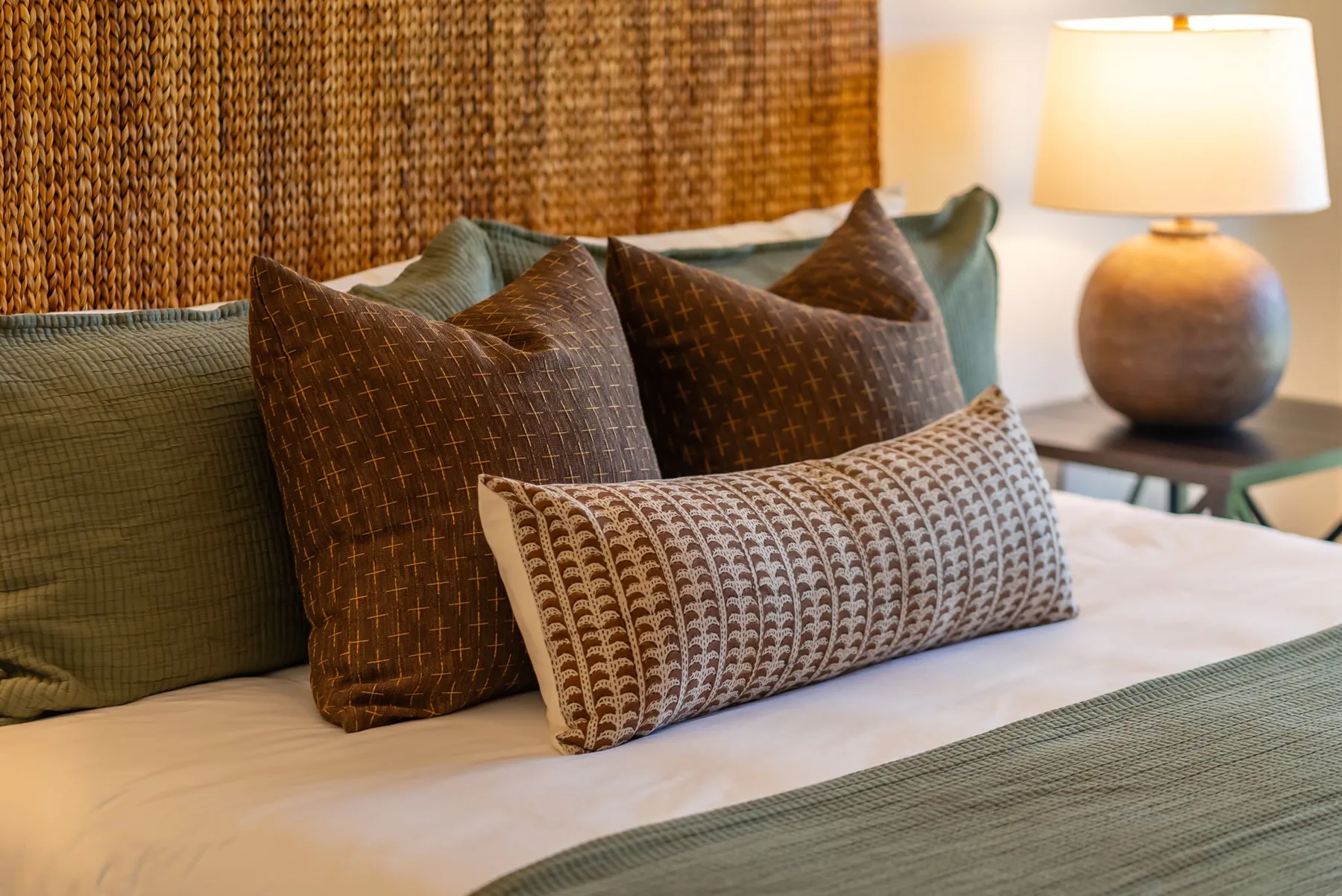 Una cama con cojines estampados en capas está junto a una lámpara de mesa encendida.