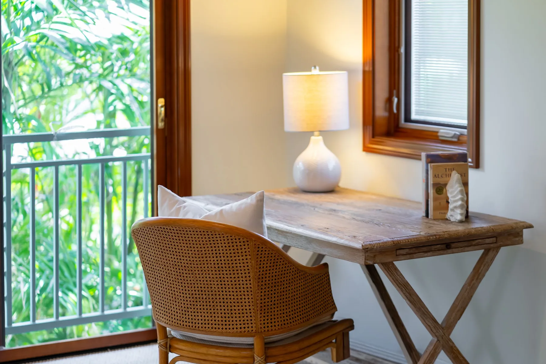 Un escritorio pequeño junto a una ventana y una puerta al balcón, con una silla y una lámpara.