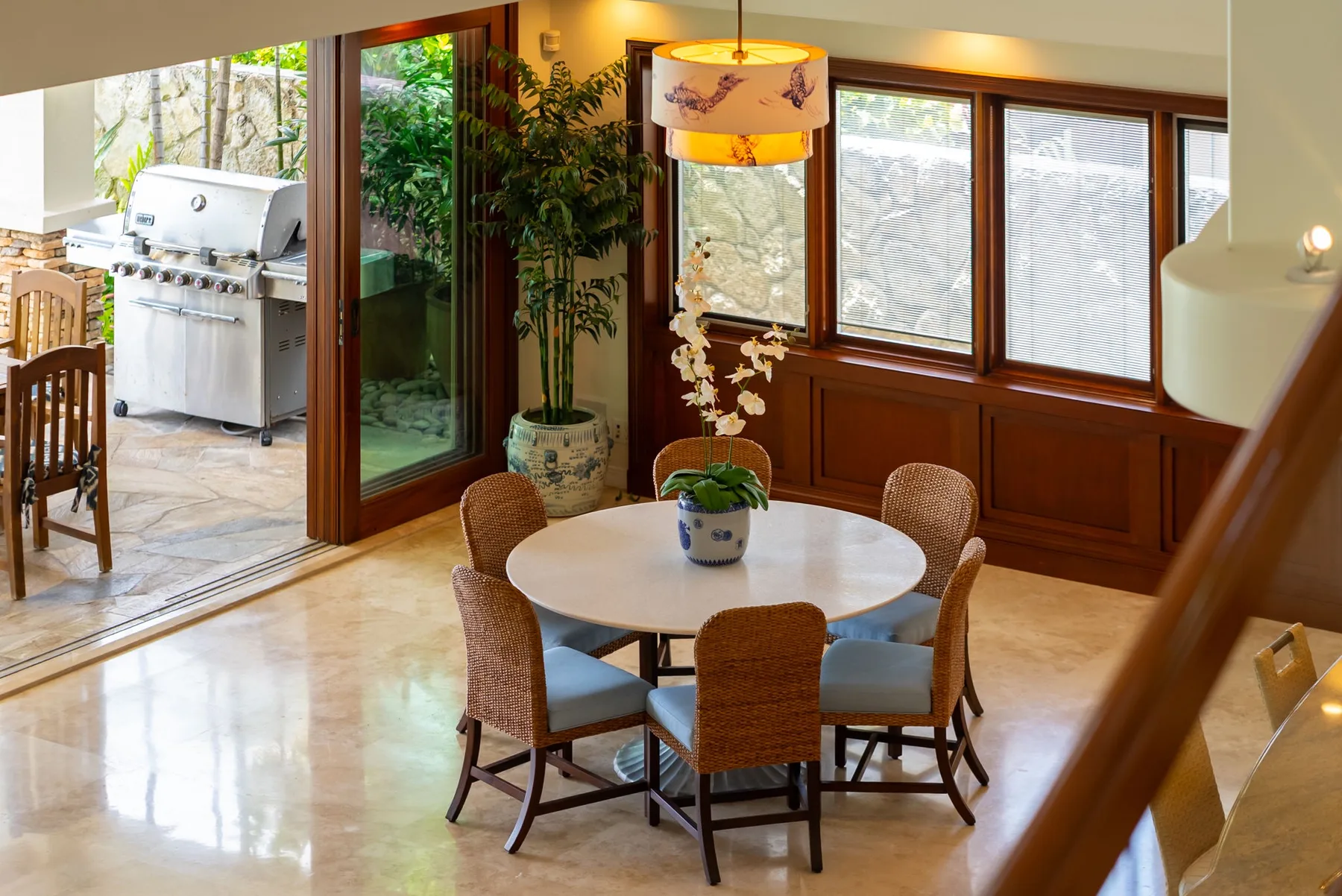 Comedor con una mesa redonda y asientos de mimbre junto a ventanas y una puerta de vidrio.