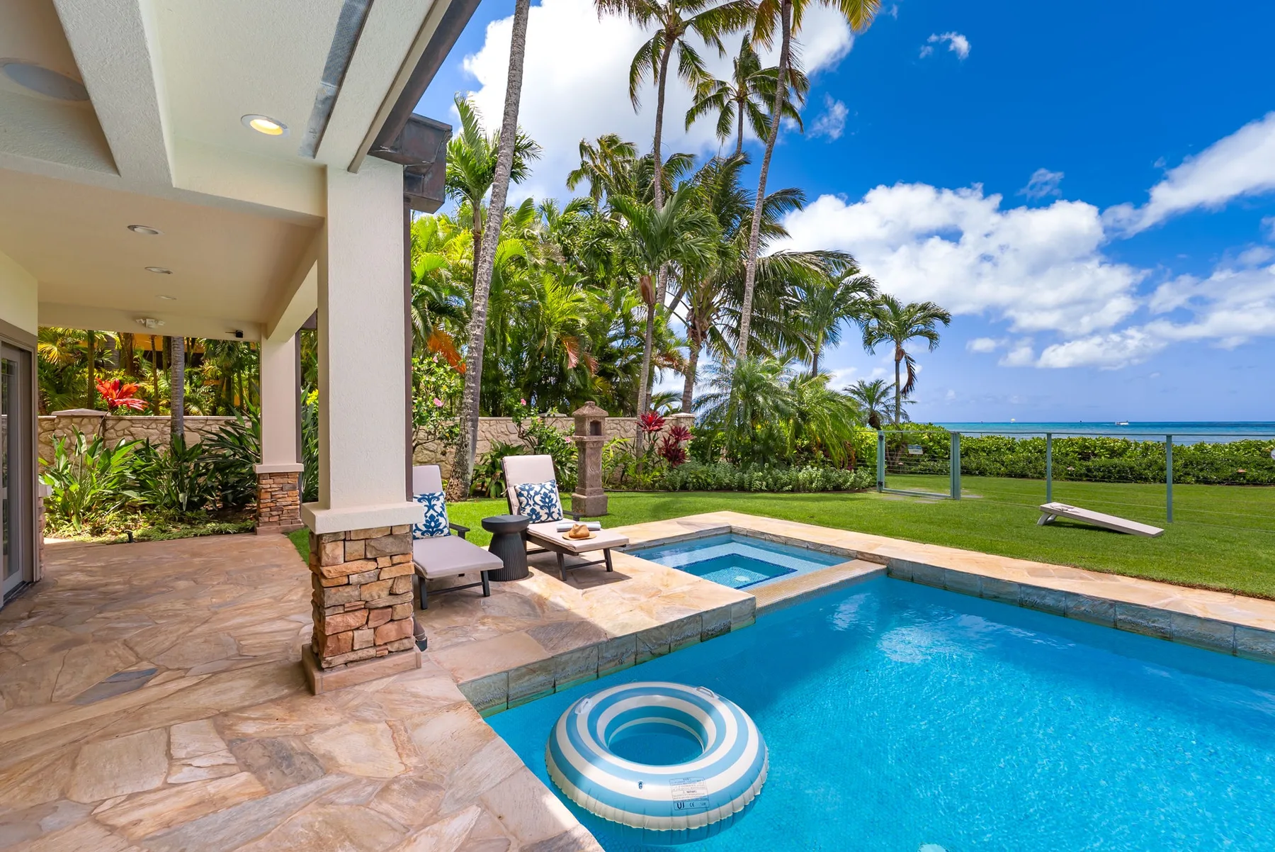 Un patio tropical da a una piscina, con palmeras y el océano al fondo.