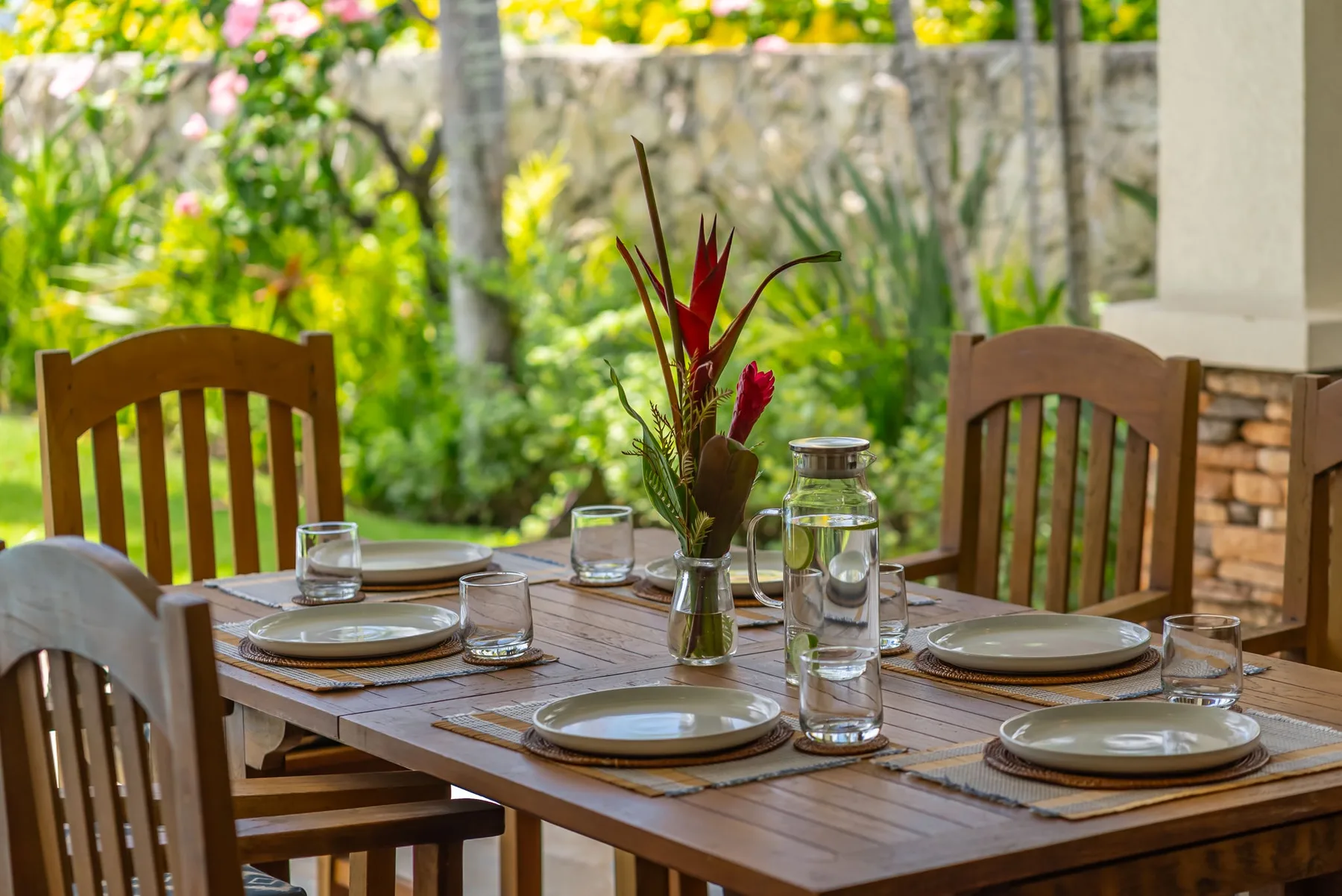 Una mesa de comedor al aire libre está puesta con platos y vasos junto a un jardín verde.