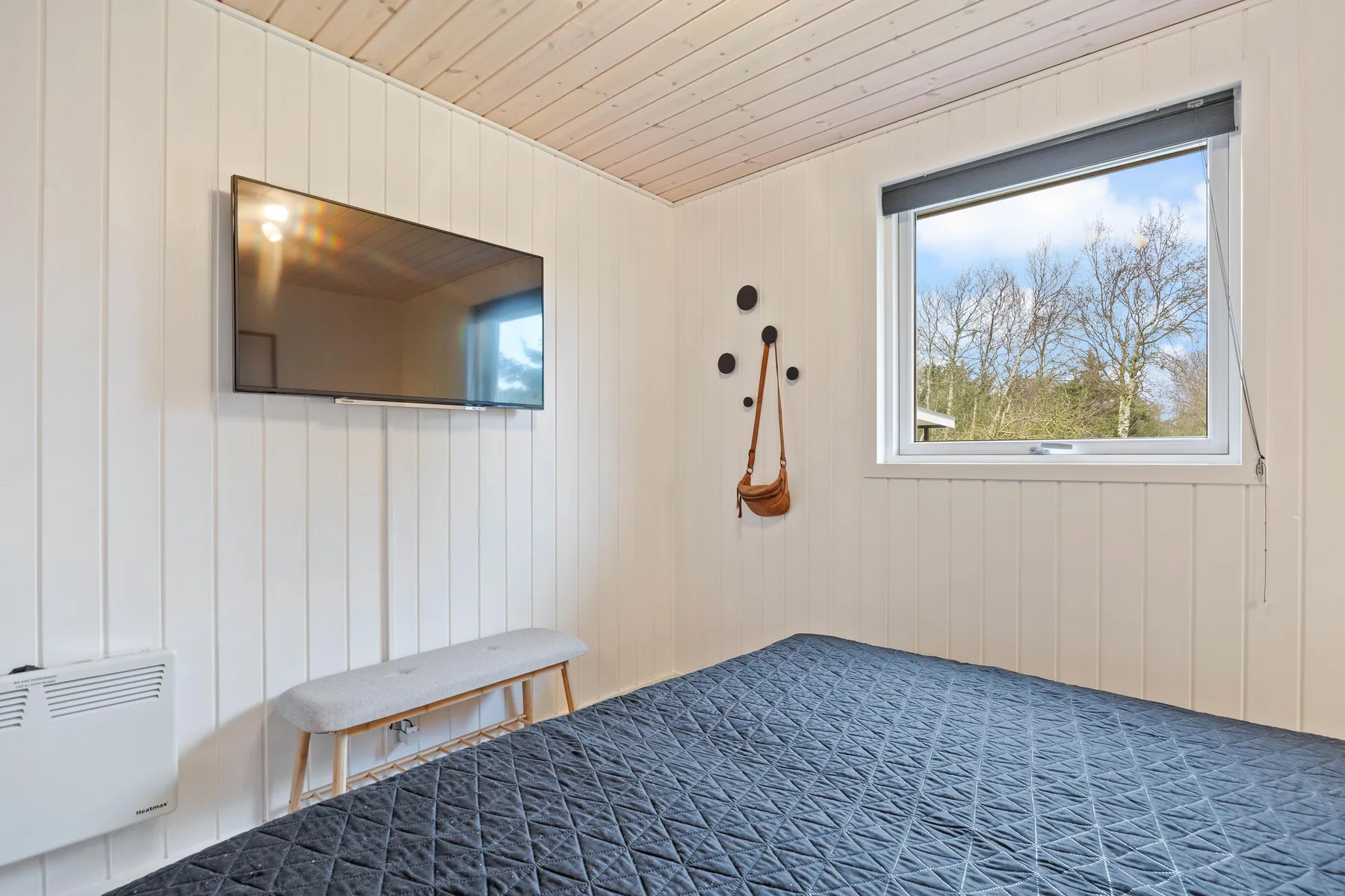 Un dormitorio luminoso con un televisor montado en la pared y una ventana con vistas a los árboles.