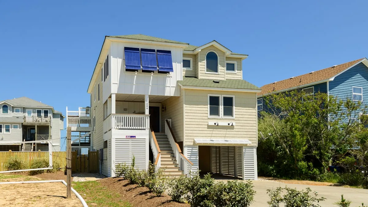 A raised coastal house with exterior stairs stands beside neighboring homes under a clear sky.