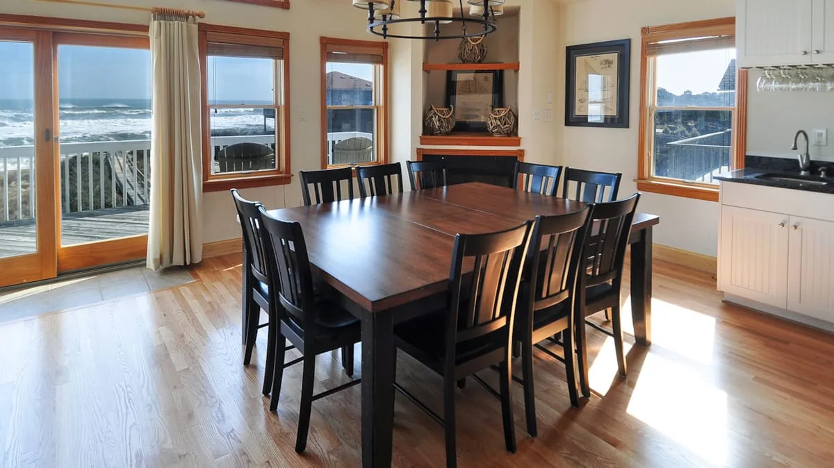 A dining area with a large wooden table and windows overlooking the water.