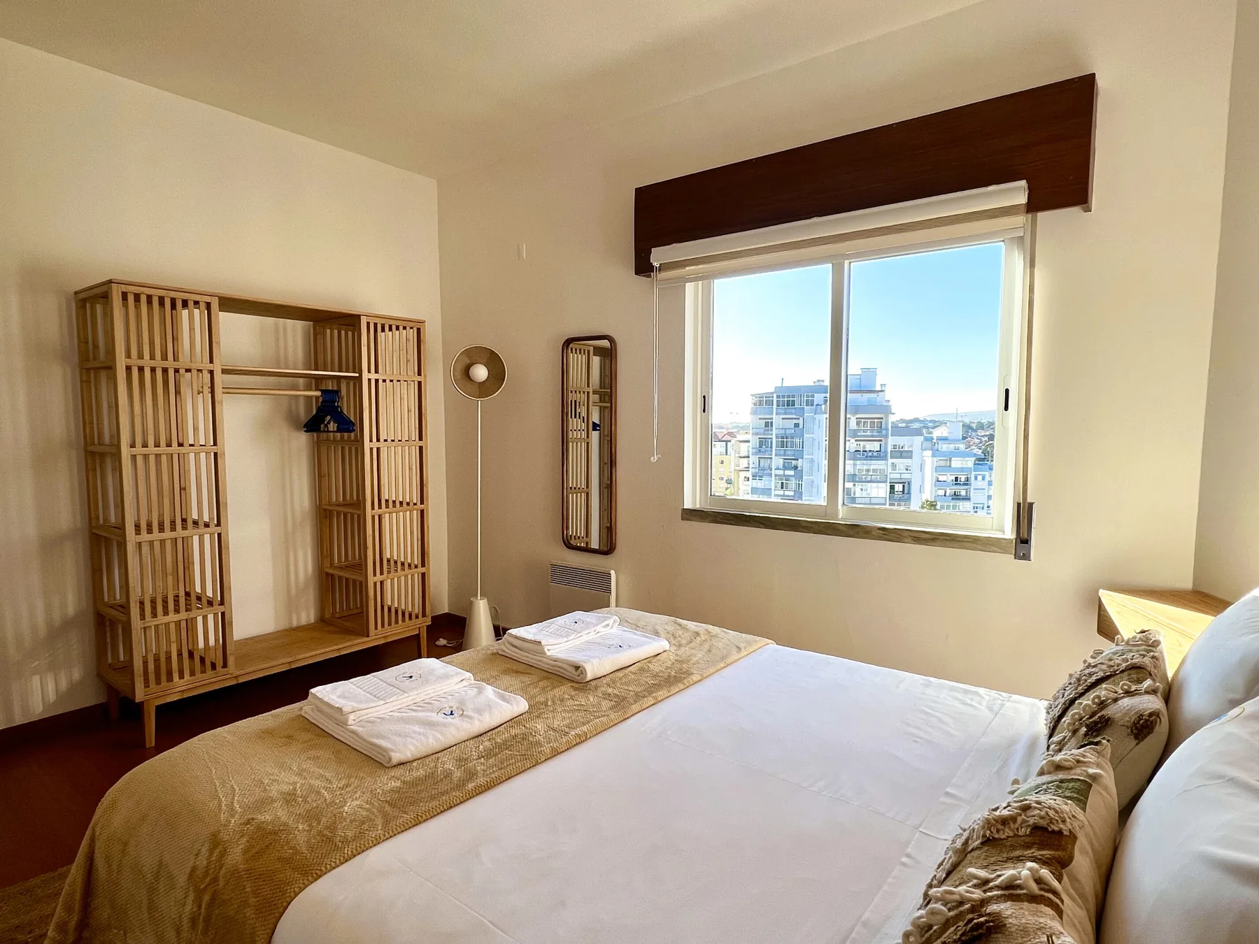 Un dormitorio luminoso con una cama hecha junto a una ventana grande con vistas a edificios cercanos.