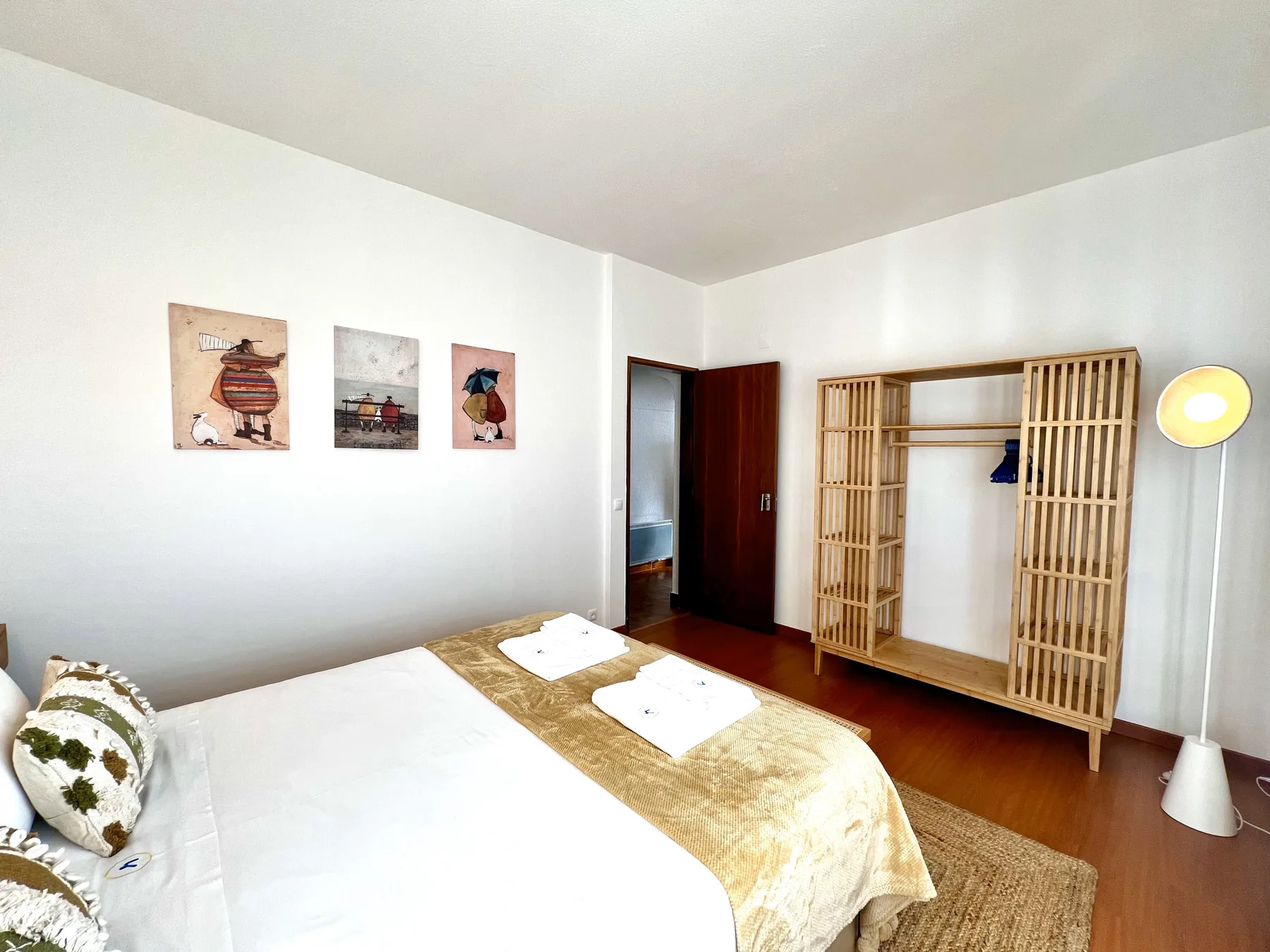 Un dormitorio luminoso con una cama, cuadros y un armario de madera abierto.