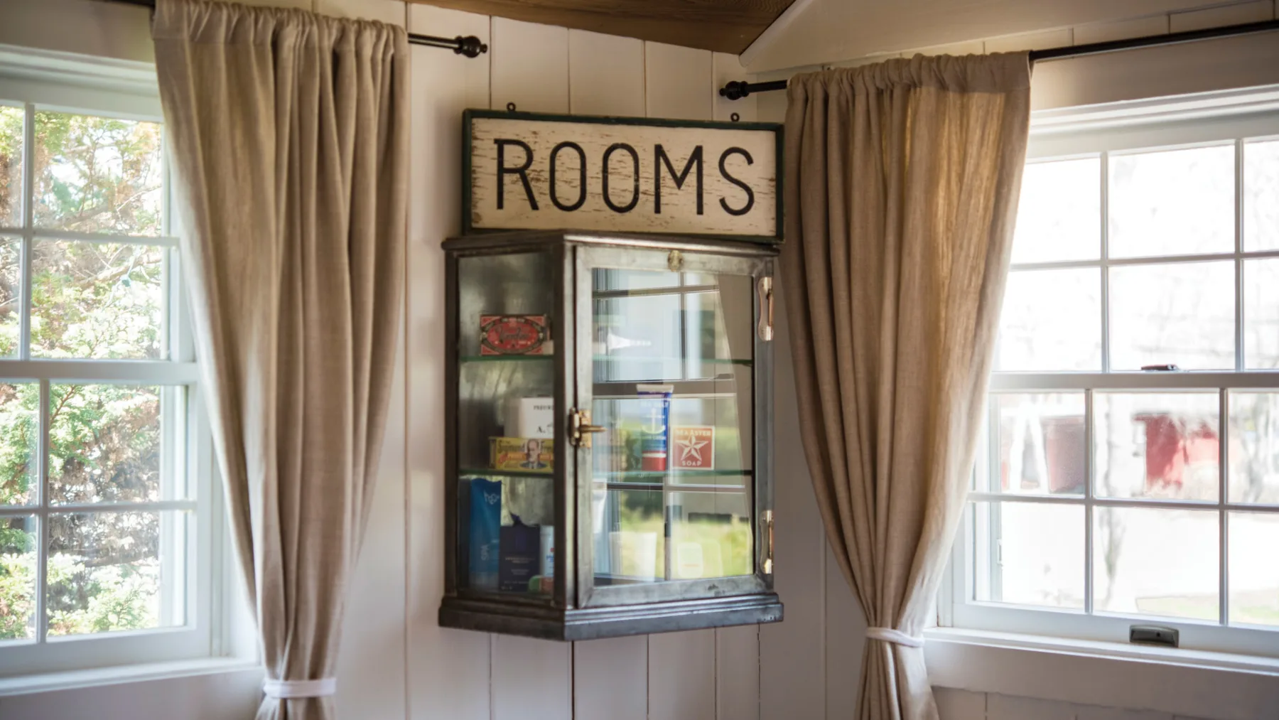 A room with large windows, curtains, and a glass-fronted cabinet labeled “ROOMS.”