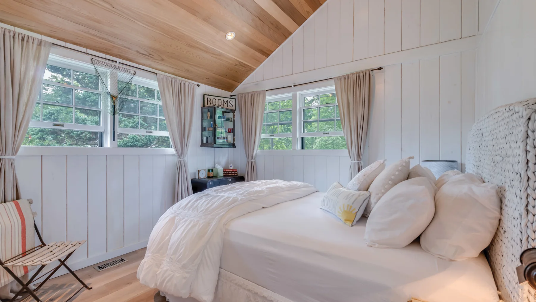 A bright bedroom with a bed beneath windows and a sloped wood ceiling.